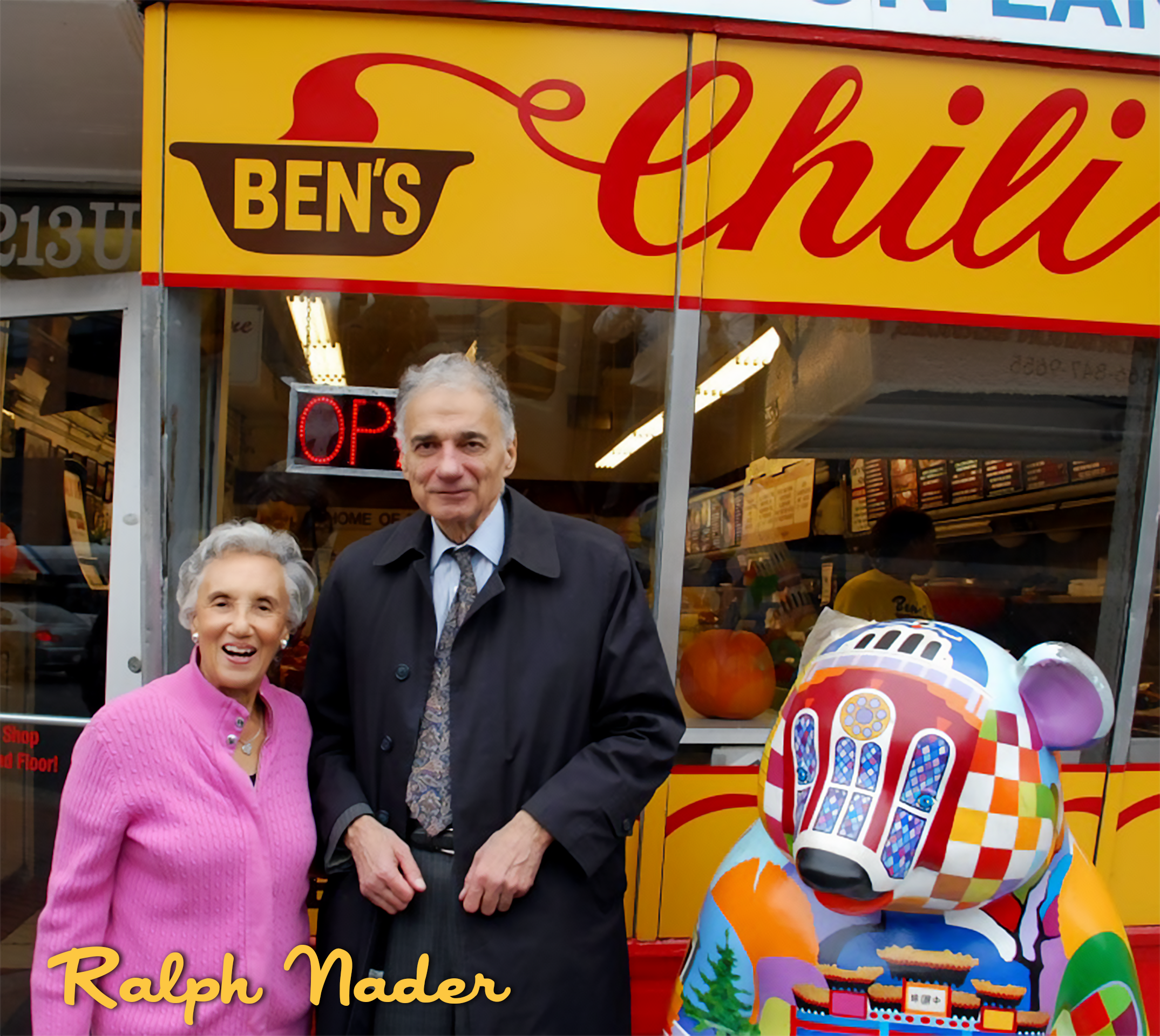 An elderly woman in a pink sweater and an elderly man in a dark coat and tie standing outside a restaurant named Ben's Chili. There is a colorful decorative sculpture of a stylized animal, possibly a bear, in front of the restaurant.
