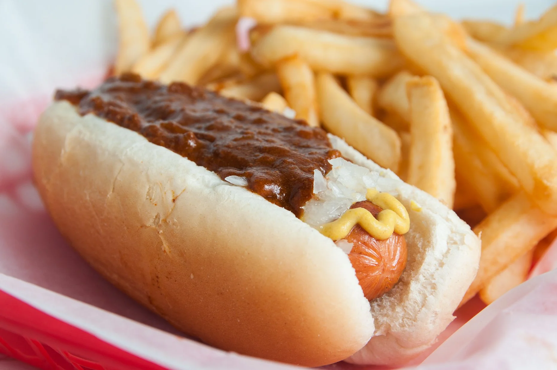 Hot dog in a bun with mustard and chili, accompanied by a side of French fries.