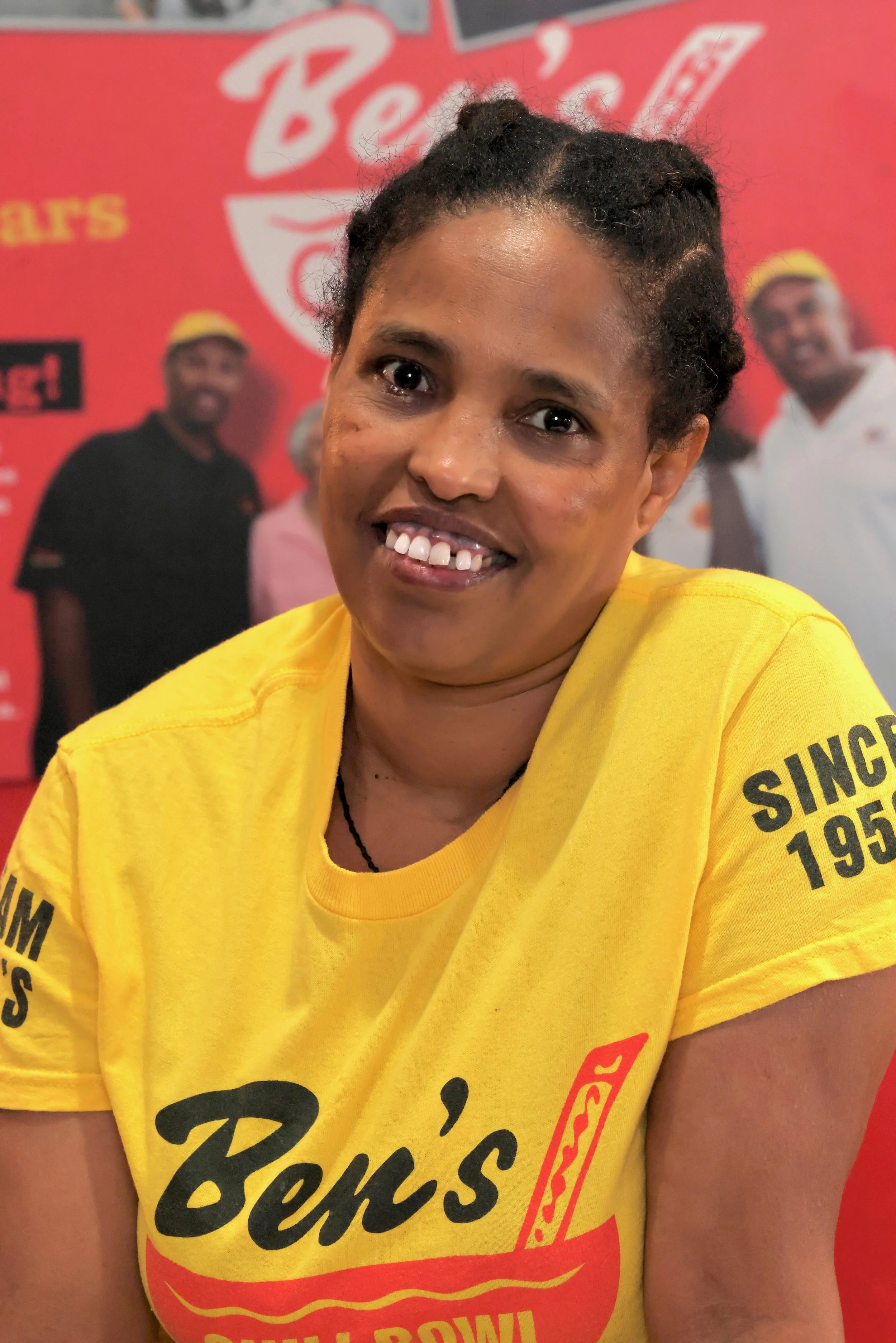 Woman in yellow Ben's chili bowl t-shirt smiling with a red Ben's chili bowl background.