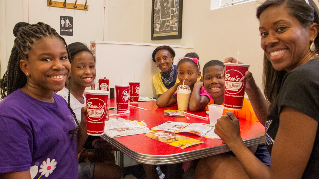 A group of six smiling children and two adults sitting around a red table at a restaurant, with drinks and menus on the table.