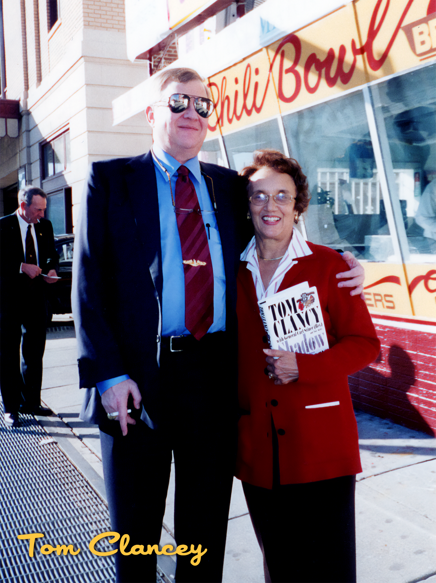 A man and woman smiling and standing together outdoors, with a bus behind them that has the words 'Phili Bowl' on it. The man is wearing sunglasses and a business suit with a red tie. The woman is holding a book titled 'Toni Clancy with her familiar 