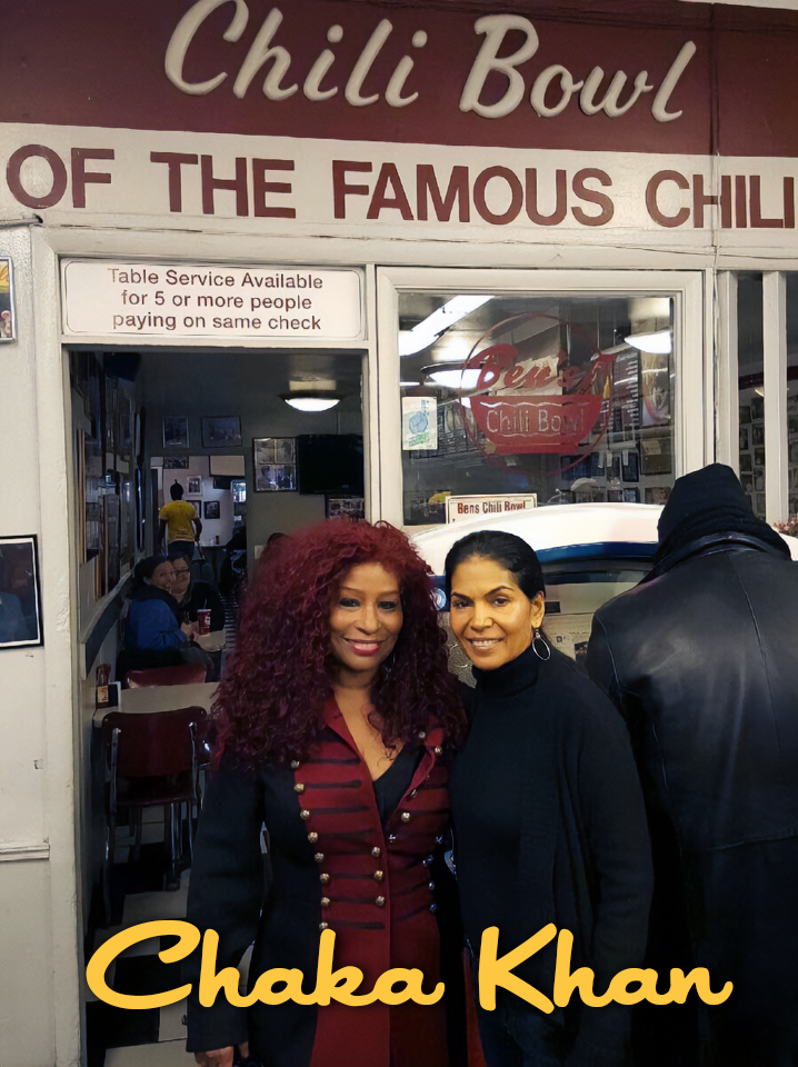 Two women smiling at the camera inside a restaurant called Chili Bowl, with a sign that says 'Chili Bowl of the Famous Chili'. They are standing outside the entrance, with a person in a leather jacket and others inside. One woman has curly red hair a
