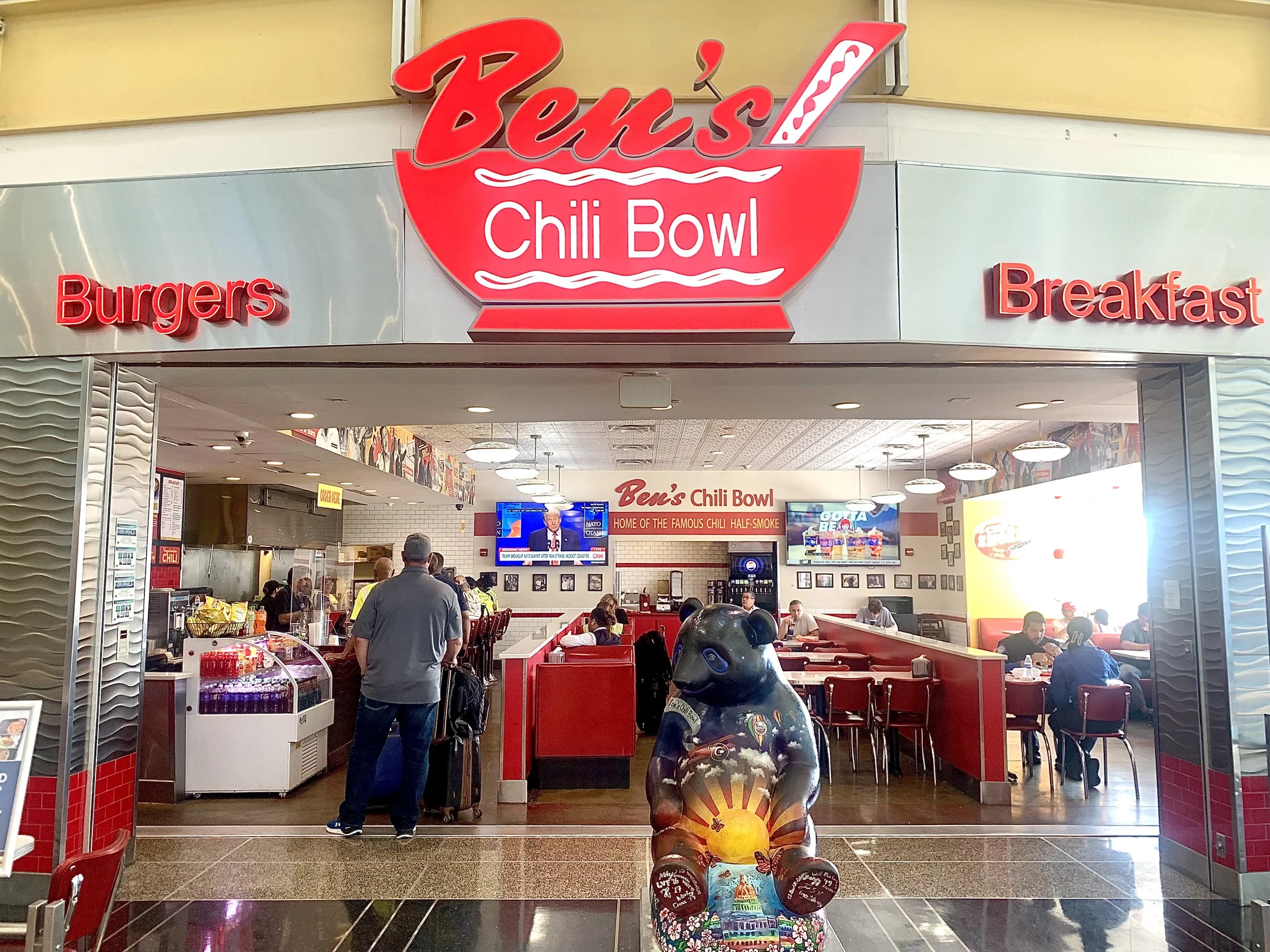 The entrance to Ben's Chili Bowl restaurant, with a bright sign featuring a red chili bowl and the restaurant's name. The entrance has a large bear statue holding a sunrise. Inside, people are seated at tables, and there are televisions on the wall.