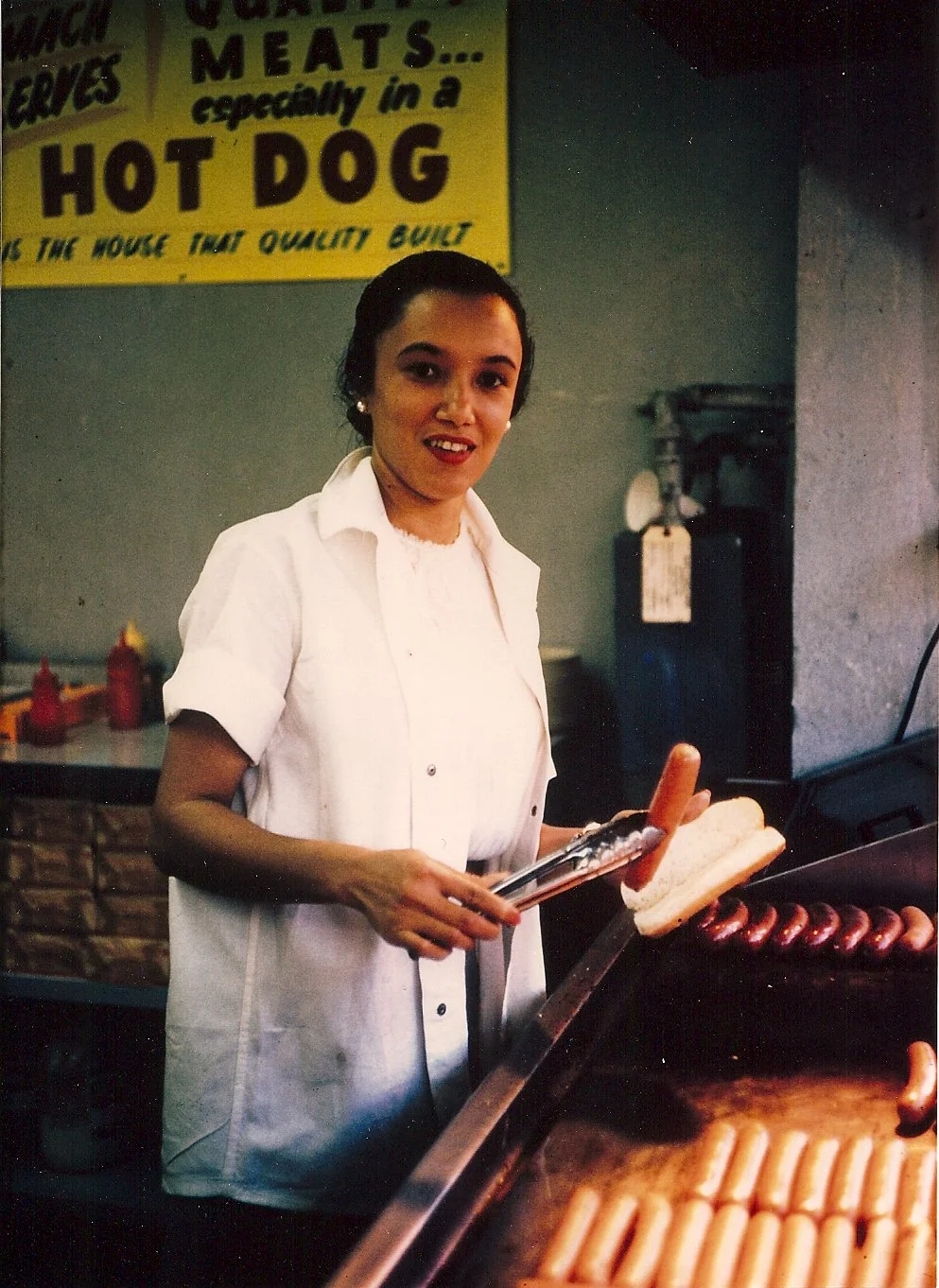 A woman grilling hot dogs at a stand, with condiments in bottles behind her and a yellow sign advertising meats and hot dogs in the background.