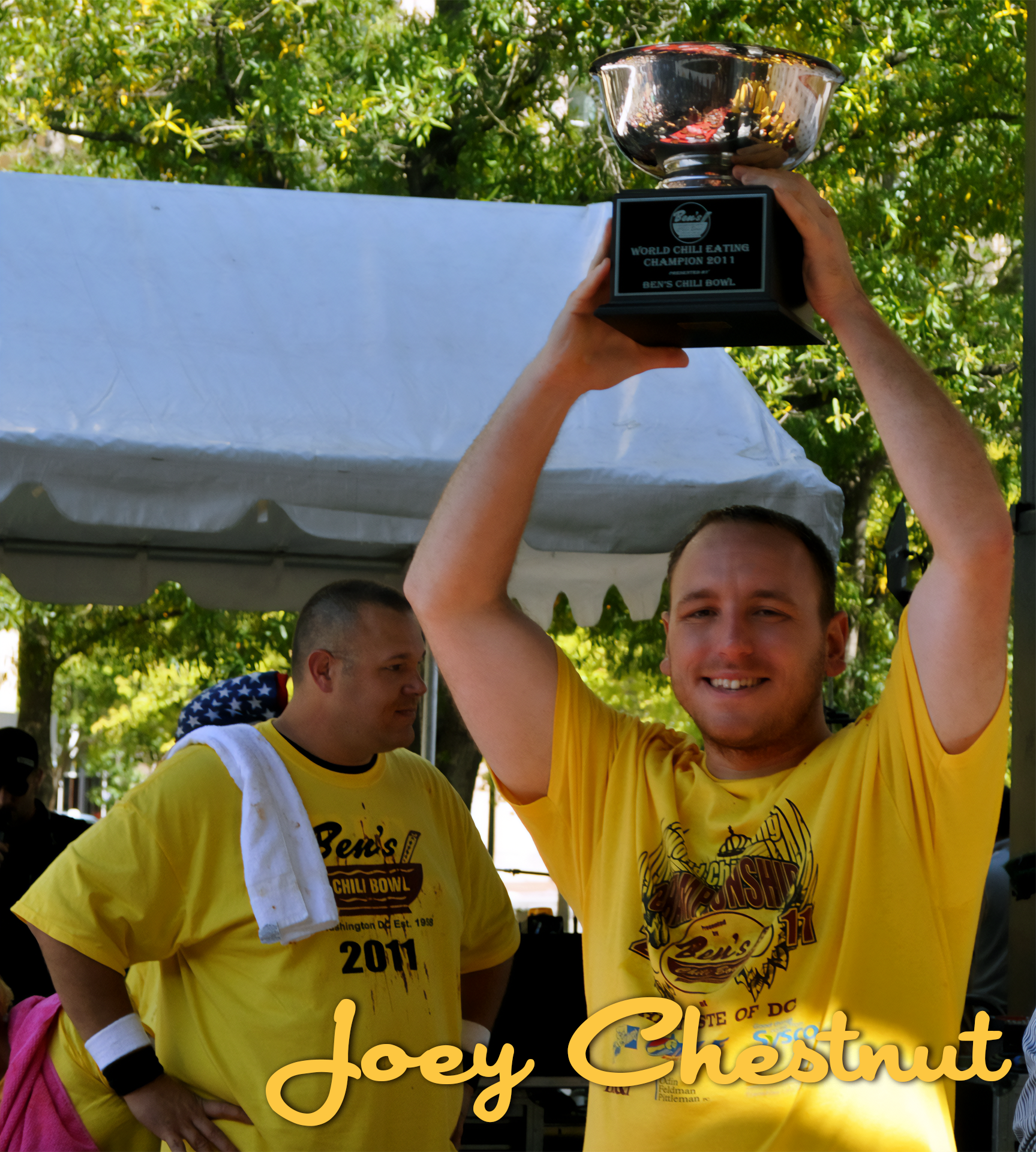 A man in a yellow t-shirt holding a trophy above his head, smiling. Another person in a yellow t-shirt stands nearby. They are outdoors under a white canopy, with trees in the background. The trophy has a plaque that reads, "World Chili Eating Champi