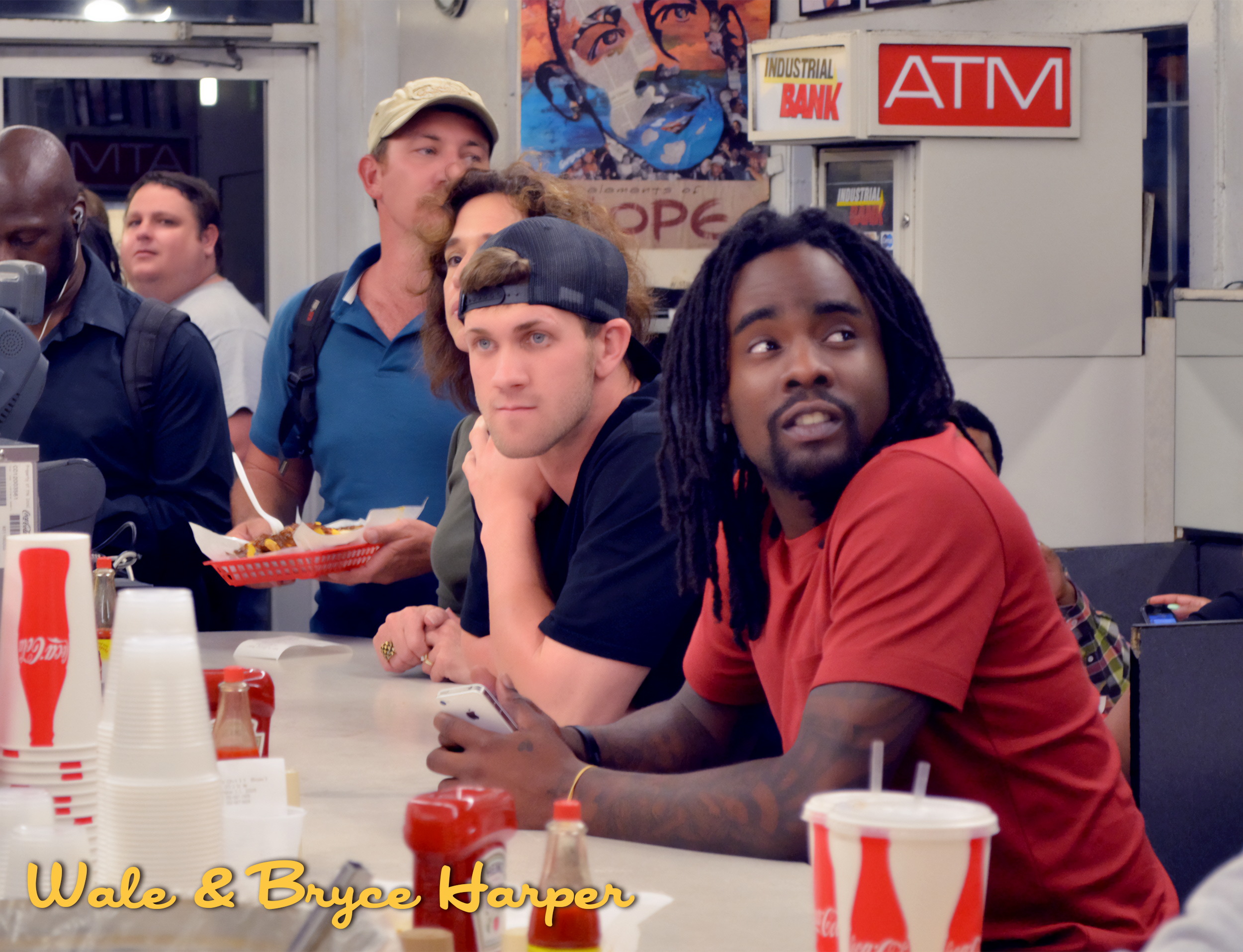 A group of people sitting at a counter inside a restaurant, with a man in a red shirt with dreadlocks holding a phone, and other people sitting and standing around, with Coca-Cola cups and condiments on the counter.