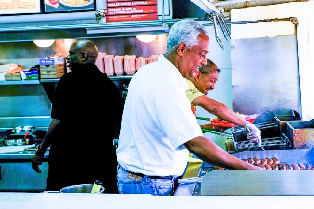 Three men cooking and working behind a counter in a restaurant kitchen. One man in a white shirt is grilling meatballs, while another in a yellow shirt and glasses is preparing food. A third man in black is looking away, near shelves with boxes and k