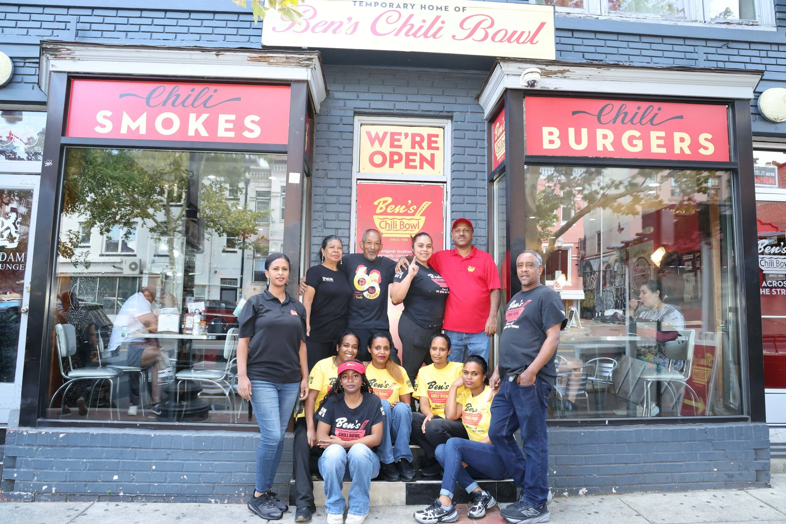 Group of people standing and sitting outside a restaurant named Ben's Chili Bowl, with signs that say 'Chili Smokes,' 'Chili Burgers,' and 'We're Open.' The restaurant has a black brick exterior with large windows.