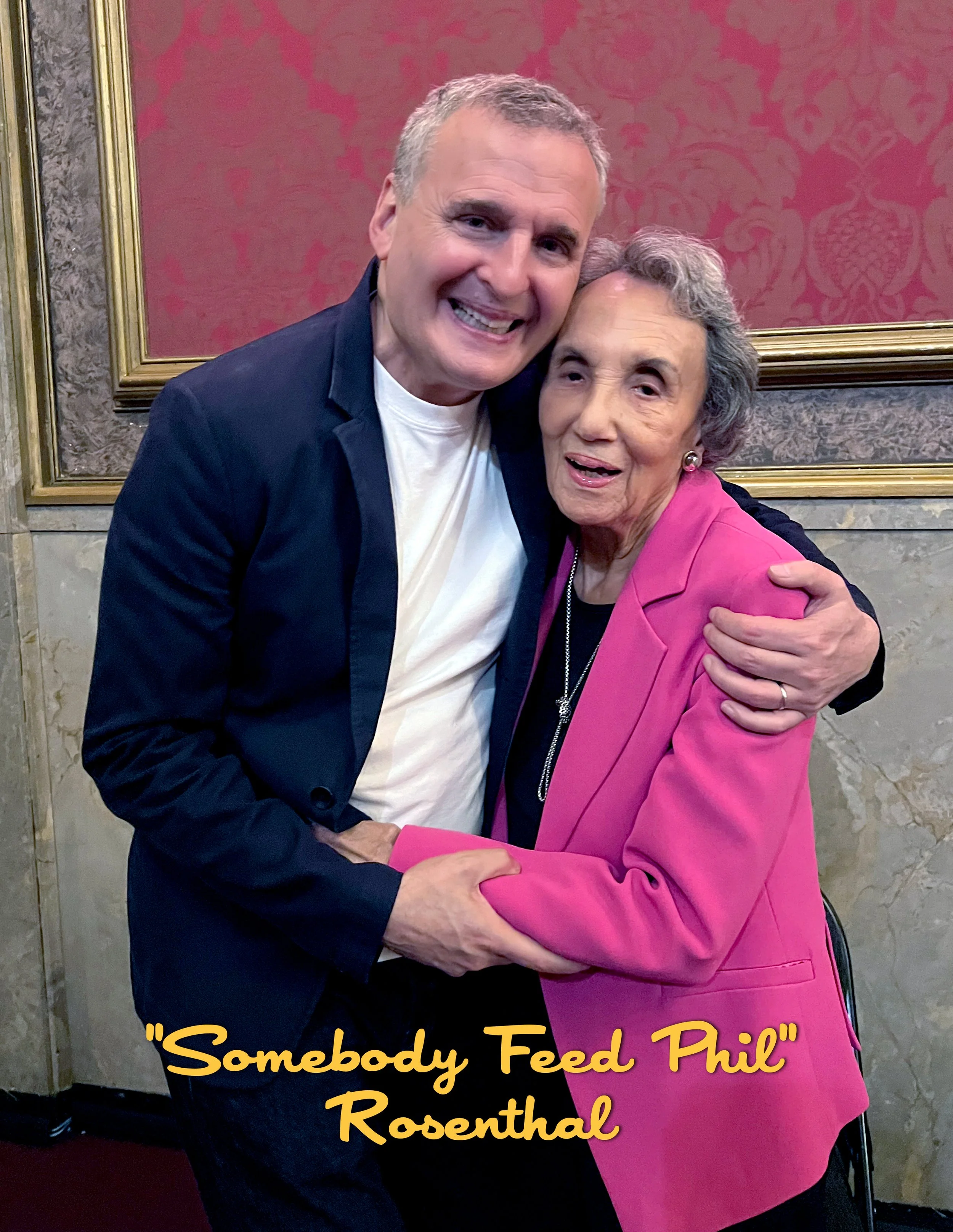 A man and an elderly woman hugging and smiling. The woman is wearing a pink blazer and the man is wearing a dark jacket and white t-shirt. There is text on the image that reads 'Somebody Feed Phil' Rosenthal.