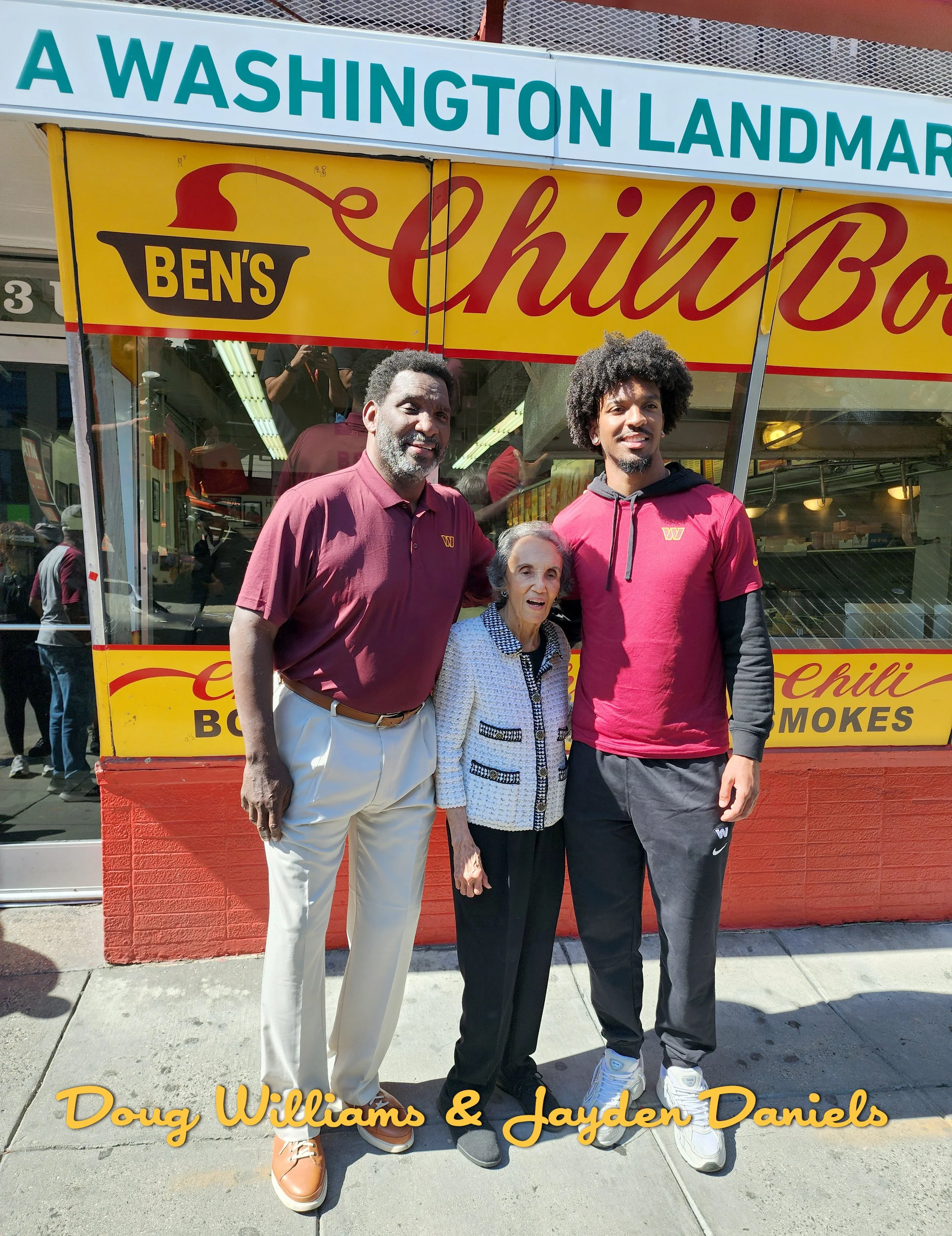 Three people, Doug Williams and Jayden Daniels, standing in front of a yellow and red food stand for Ben's Chili Bowl, with a sign above that says 'A Washington Landmark.'