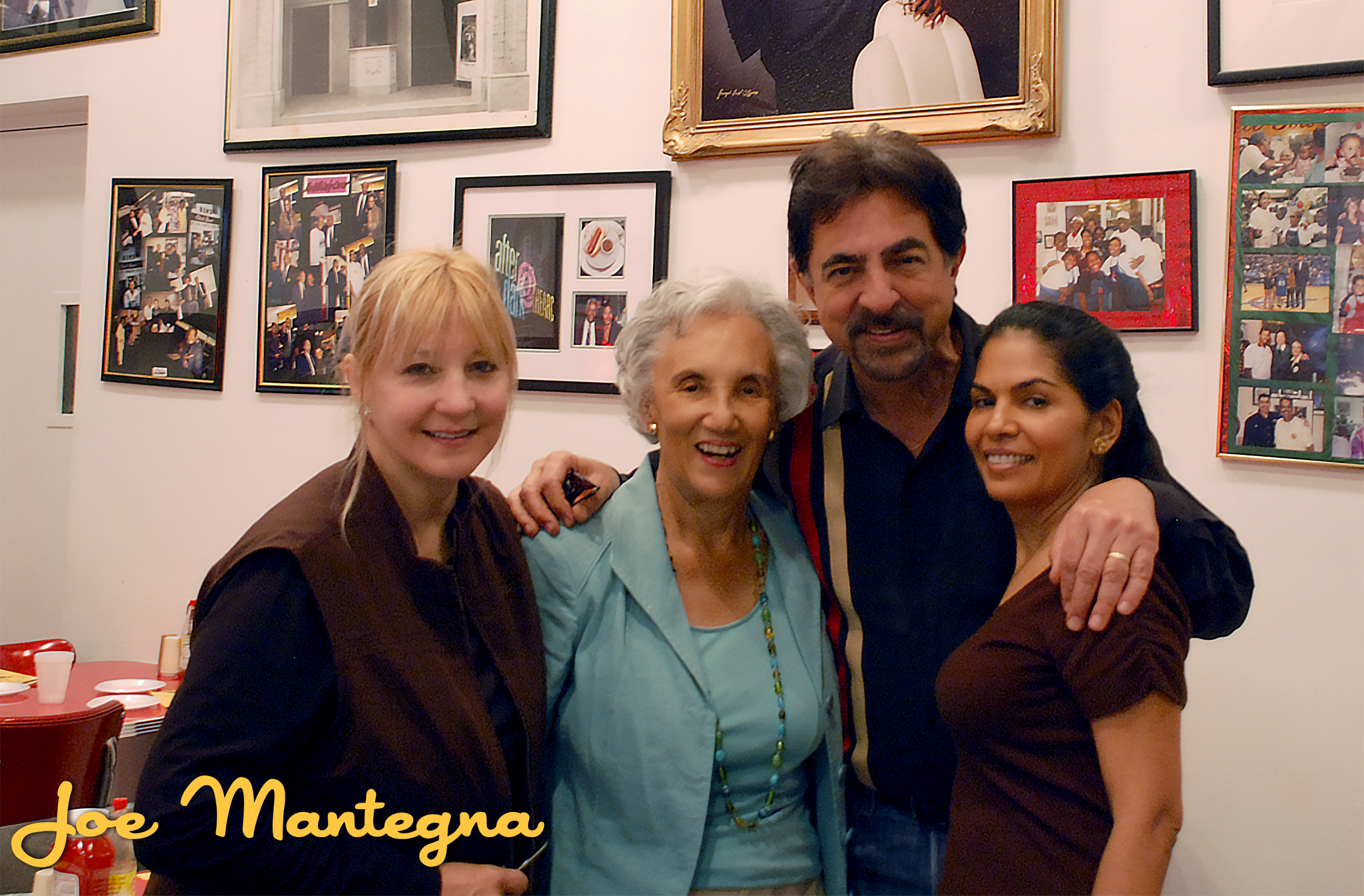 Four people standing together in front of a wall decorated with framed photographs. The group includes three women and one man, all smiling. The woman on the left has light hair and is wearing a dark brown and black outfit. The elderly woman in the c