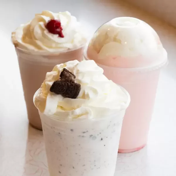 Three milkshakes topped with whipped cream, one with a cherry, one with chocolate piece, and one plain, on a white surface.