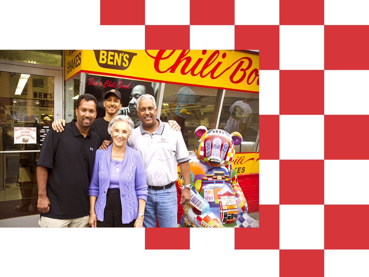 Four people standing outside a colorful food truck named Ben's Chili Bowl, with a mascot statue of a colorful, blocky bear in front.