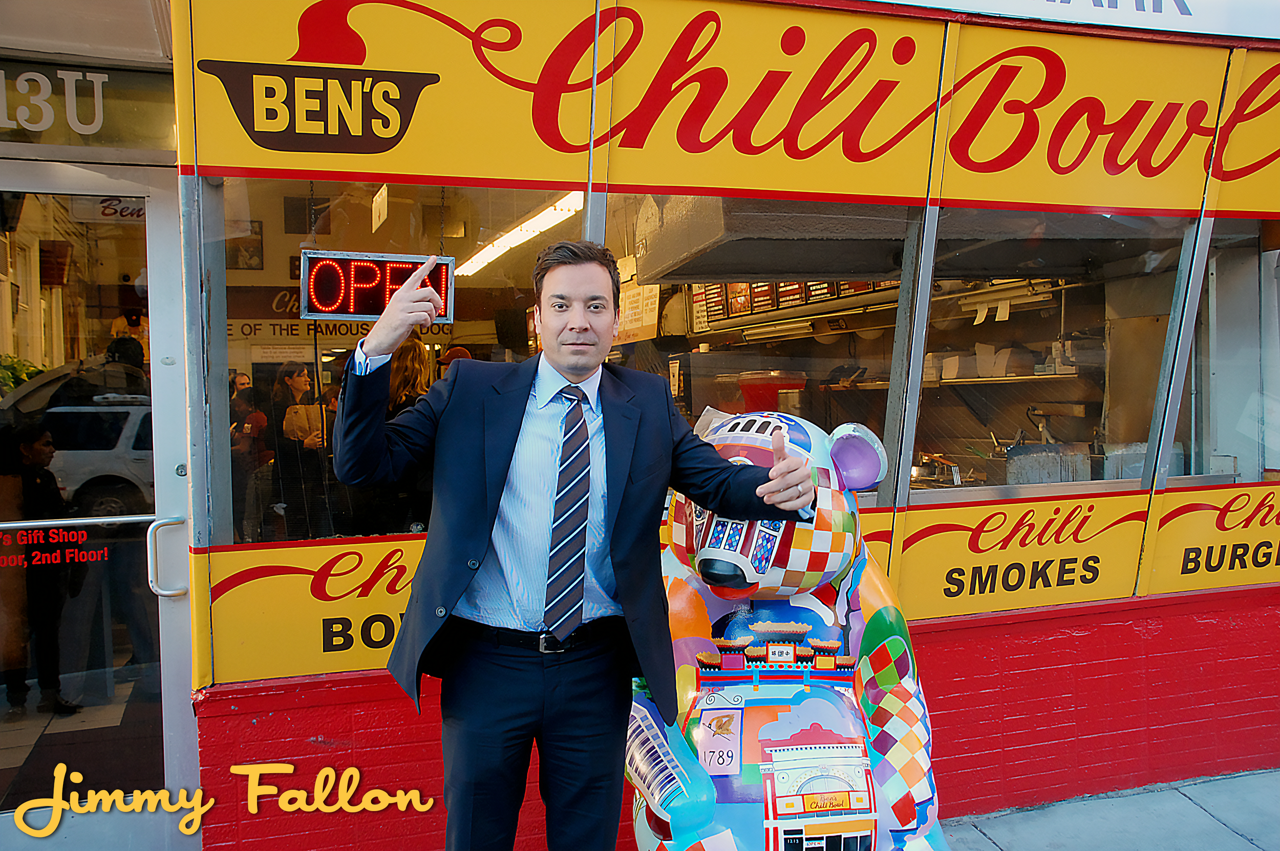 A man in a suit stands in front of a colorful food stand with a large painted bear. He points upward with one finger and gives a thumbs-up with the other. The stand is named Ben's Chili Bowl and features a yellow and red sign. Behind him, inside the 