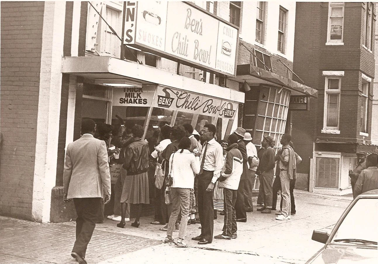 Black and white photo of a group of people standing in line outside Ben's Chili Bowl on a city street, with brick buildings in the background.