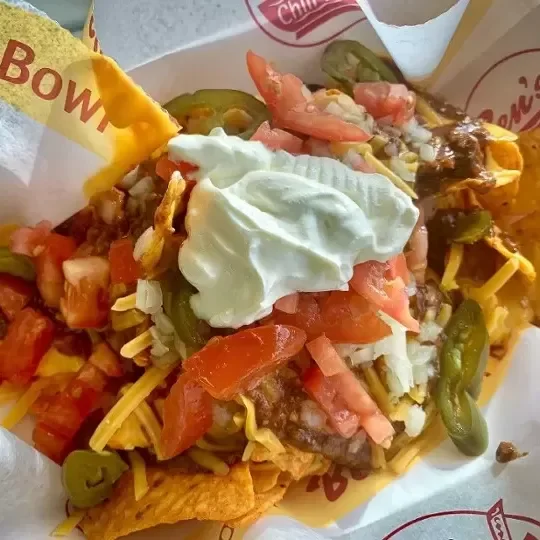 Close-up of a taco topped with sour cream, diced tomatoes, jalapeños, shredded cheese, and ground beef, served in a paper taco holder.