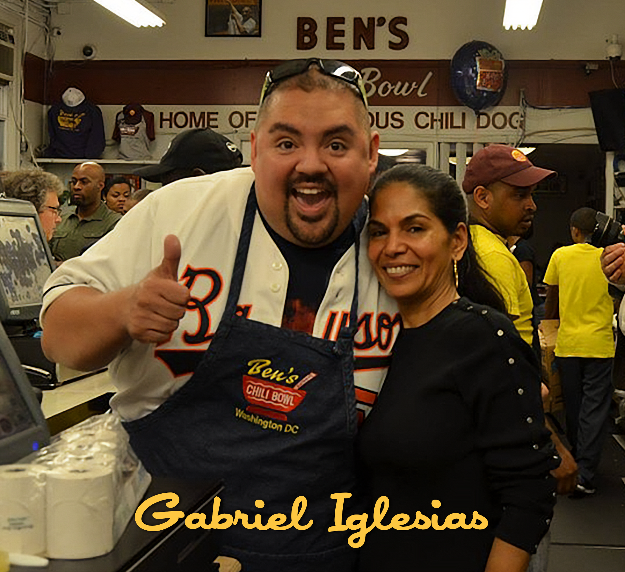 A man and woman smiling and posing together inside Ben's Chili Bowl restaurant, with the man wearing a Ben's Chili Bowl apron and giving a thumbs up. The background shows other customers and the restaurant's signage.