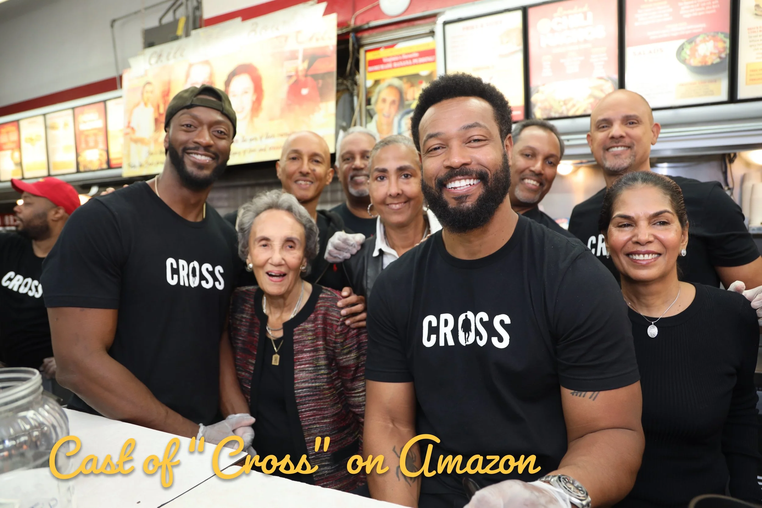 Group of people dressed in black "Cross" t-shirts gathered inside a fast food restaurant, smiling at the camera. The background shows menu boards with food options, and an elderly woman standing in the middle is surrounded by others. The text overlay