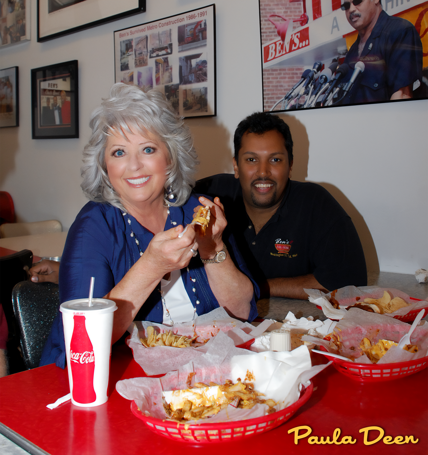 A smiling elderly woman with gray hair holding food, sitting at a table with a younger man with dark hair. They are in a casual dining restaurant, with food and drinks on the table. On the wall behind them are framed photos and a large screen display