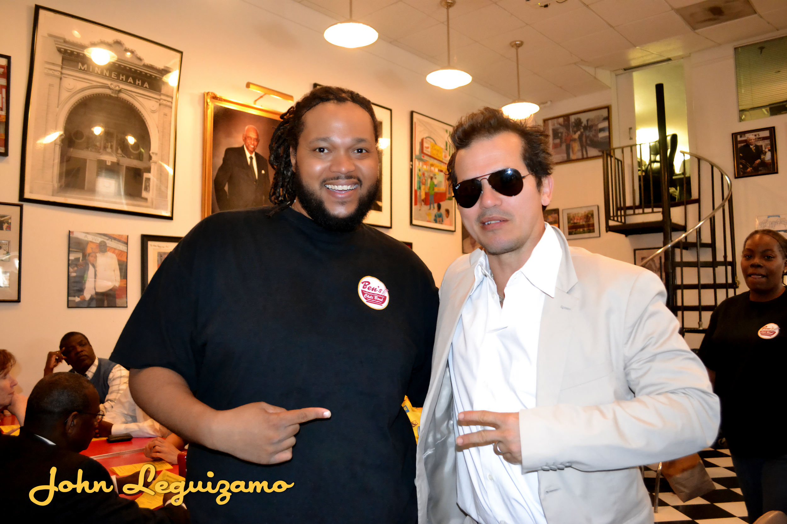 A man with dreadlocks and a beard wearing a black Ben's Chili Bowl t-shirt is smiling and pointing at a man in sunglasses and a white blazer. They are in a room decorated with framed photographs and artwork, with a staircase in the background.