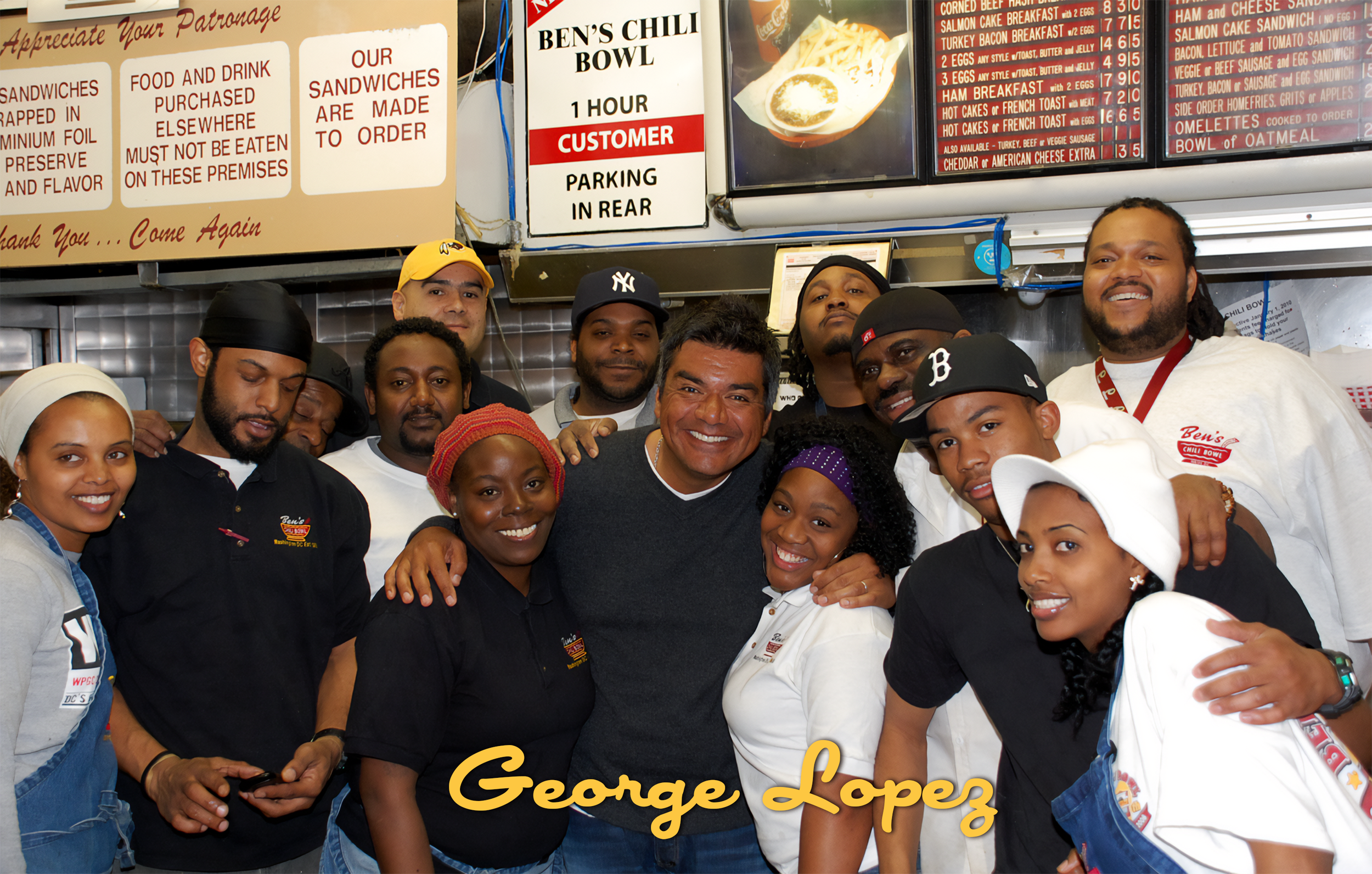 A group of smiling people, including George Lopez, in a restaurant or food stand setting with menu boards overhead, some wearing uniforms or aprons, posing for a photo.