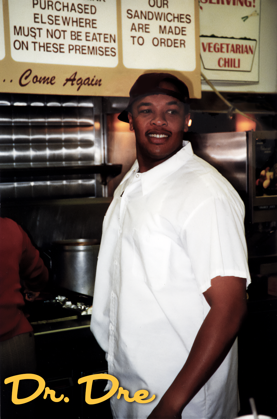 A young man wearing a white shirt and black cap, smiling in a restaurant or diner. Behind him are signs with text about ordering sandwiches, vegetarian chili, and a note about where purchased items cannot be eaten. The text 'Dr. Dre' is written in ye