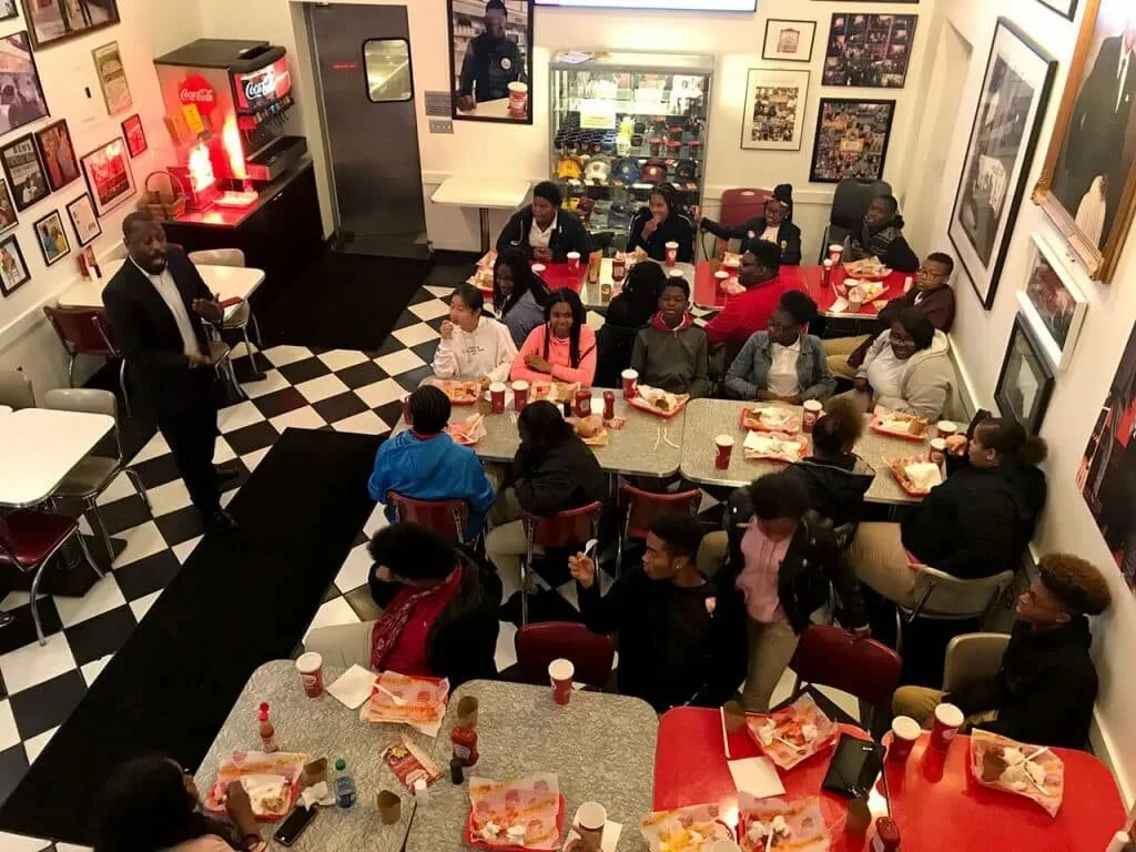 Group of people sitting around dining tables in a pizza restaurant, listening to a man in a suit speaking.