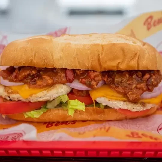 Close-up of a loaded chicken sandwich with fried chicken, lettuce, tomato, onion, cheese, and sauce in a sandwich bun, served with fries wrapped in a paper