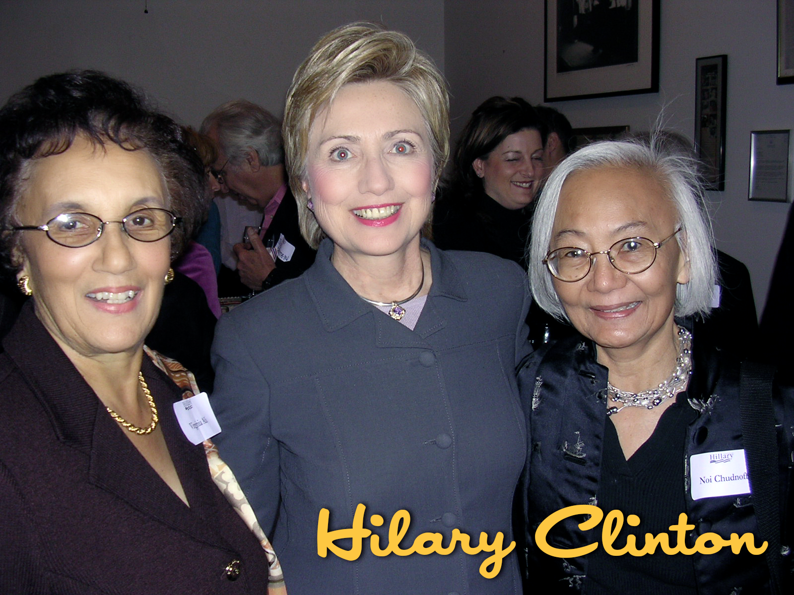 A group of women smiling at an indoor event, with a woman in the center who is likely Hillary Clinton, wearing a dark jacket. Other women around her have glasses and name tags. The background shows people and framed pictures on the wall.