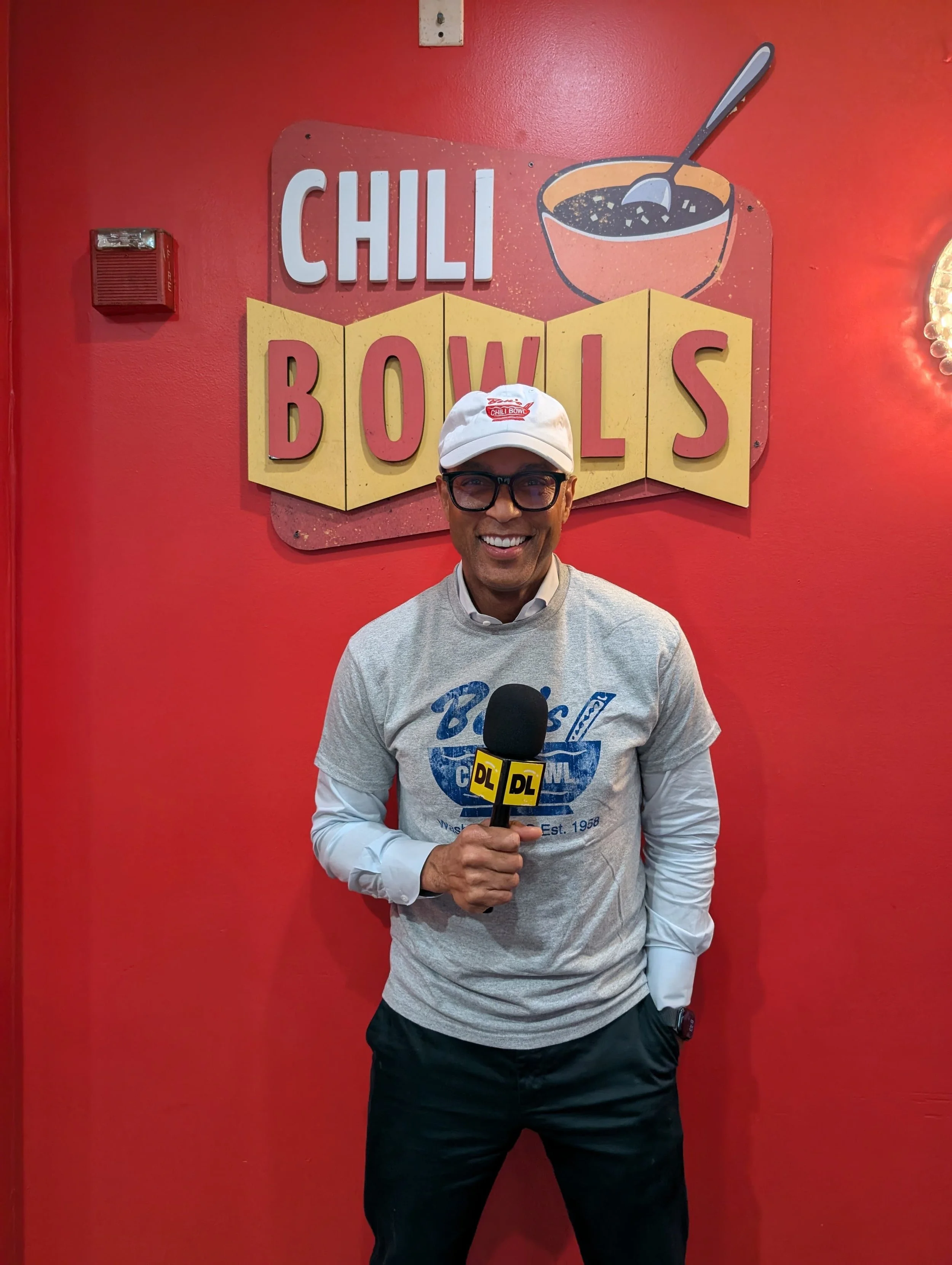 A man smiling, wearing glasses, a light gray shirt with a blue and white logo, a white hoodie, black pants, and a white cap with a red logo. He is holding a microphone with a yellow 