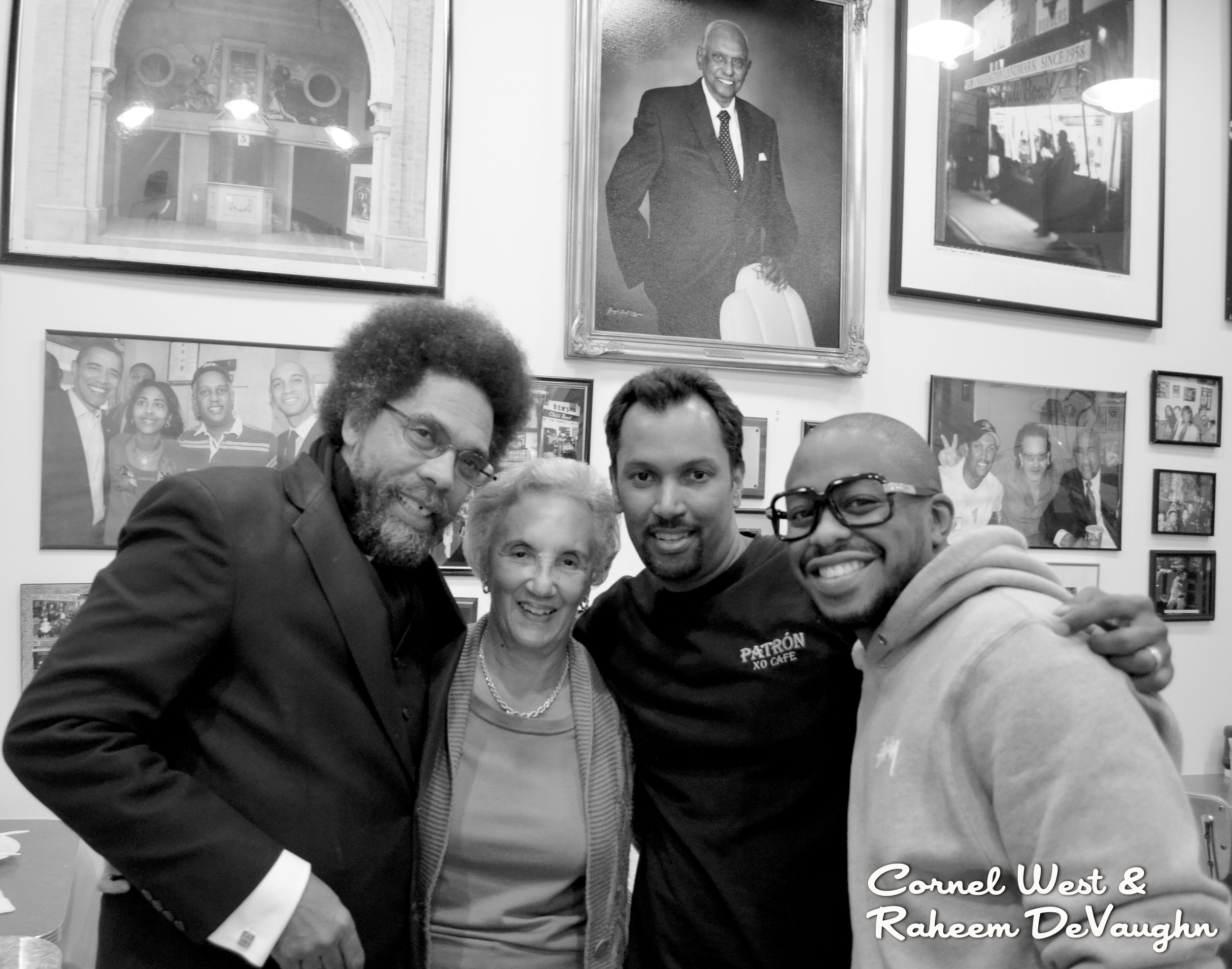 Four people standing close together in a social setting, with framed photographs and portraits on the wall in the background. Title text at the bottom right reads "Cornel West & Raheem DeVaughn."