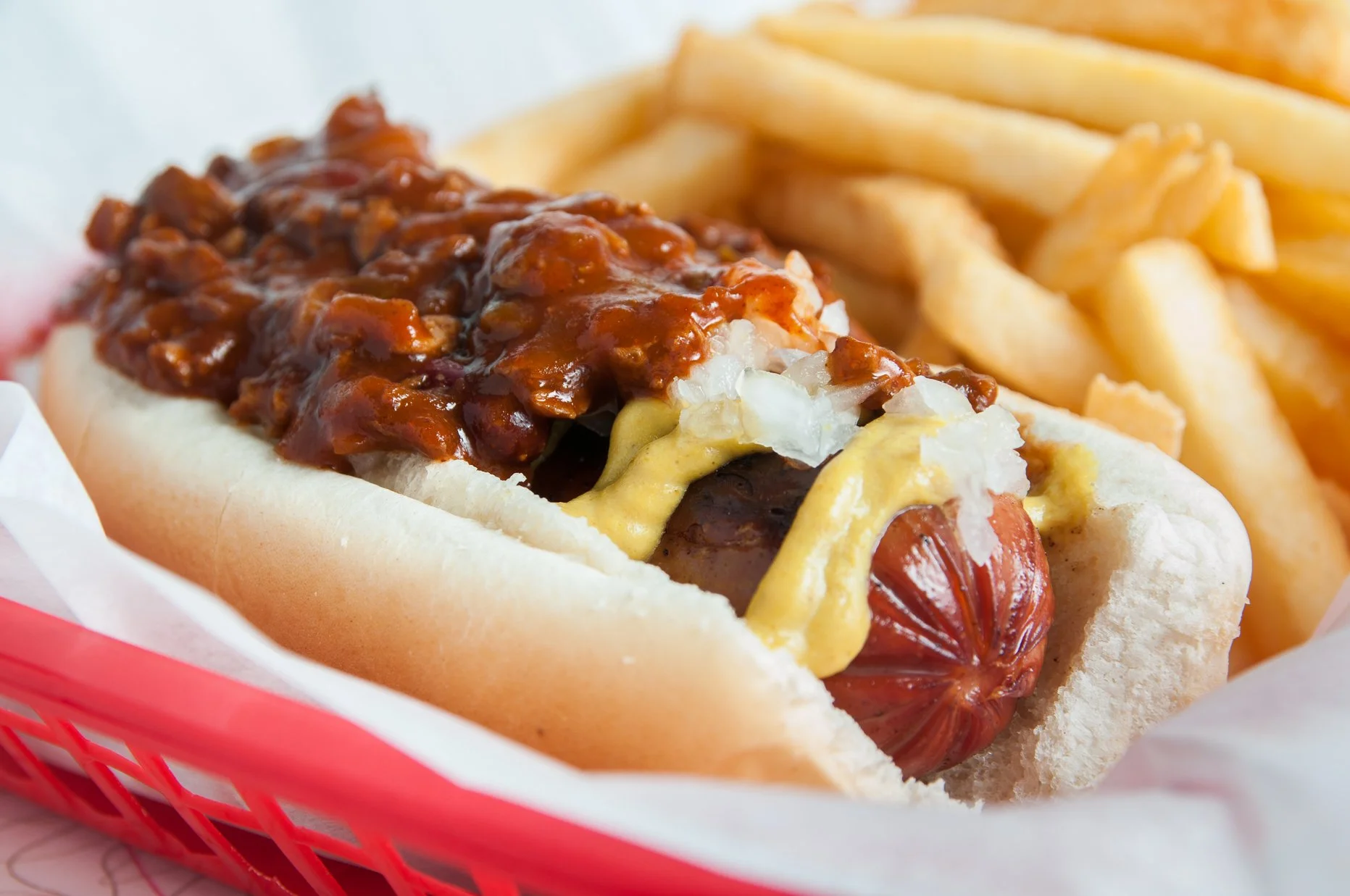 Hot dog in a bun topped with chili, mustard, and onions, served with a side of French fries in a red basket