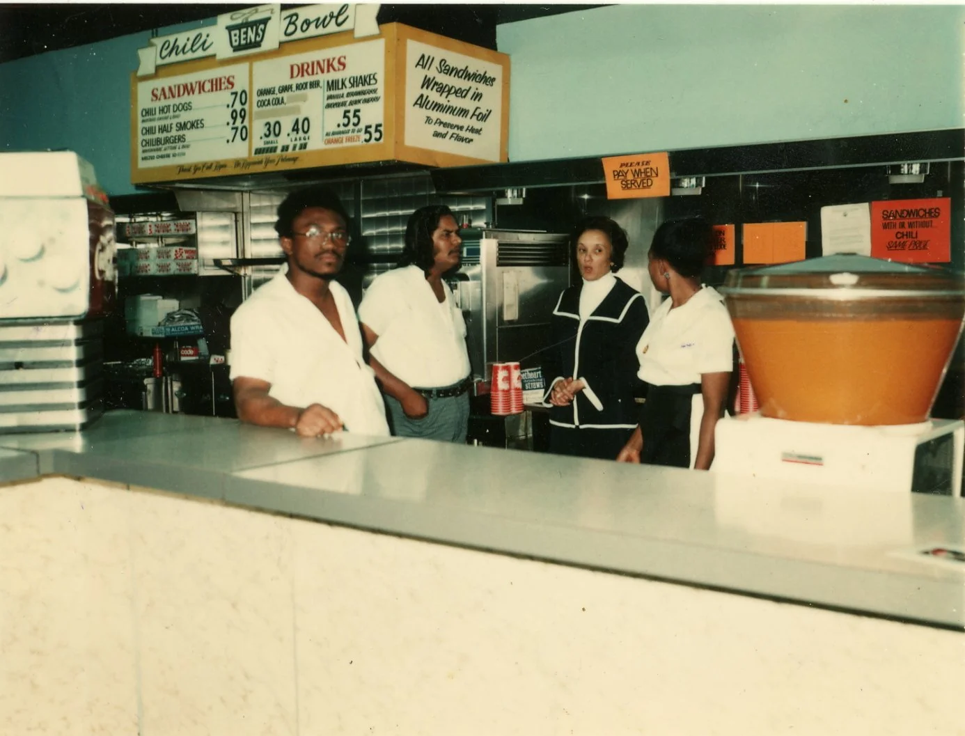 Four people, three women and one man, standing behind a counter at a fast-food restaurant named Chili Bens, with menu signs in the background.