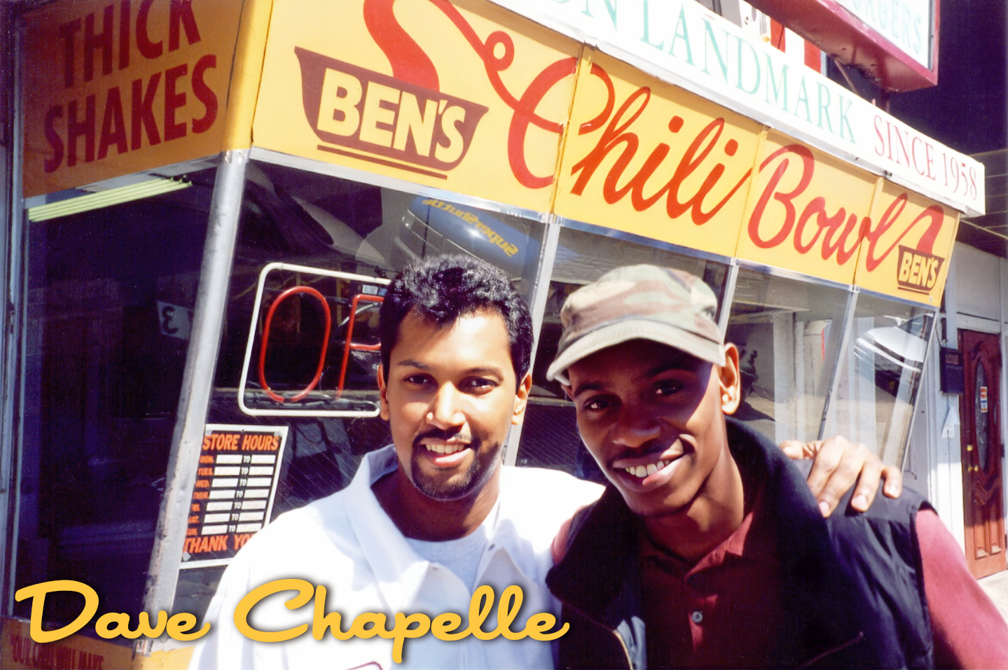 Two men standing in front of a Ben's Chili Bowl restaurant, smiling at the camera. The restaurant sign is visible in the background.