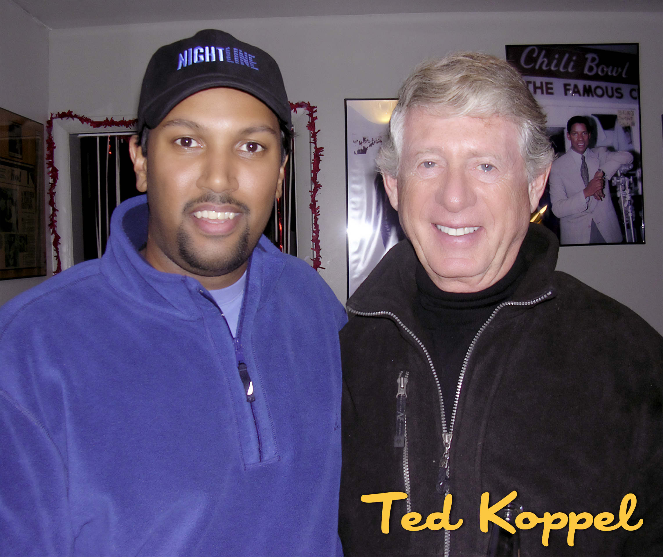 Two men standing close together, indoors, smiling for the camera. The man on the left wears a black cap and a blue zip-up jacket, and the man on the right has gray hair and wears a black zip-up jacket. A framed photograph and a poster are visible on 