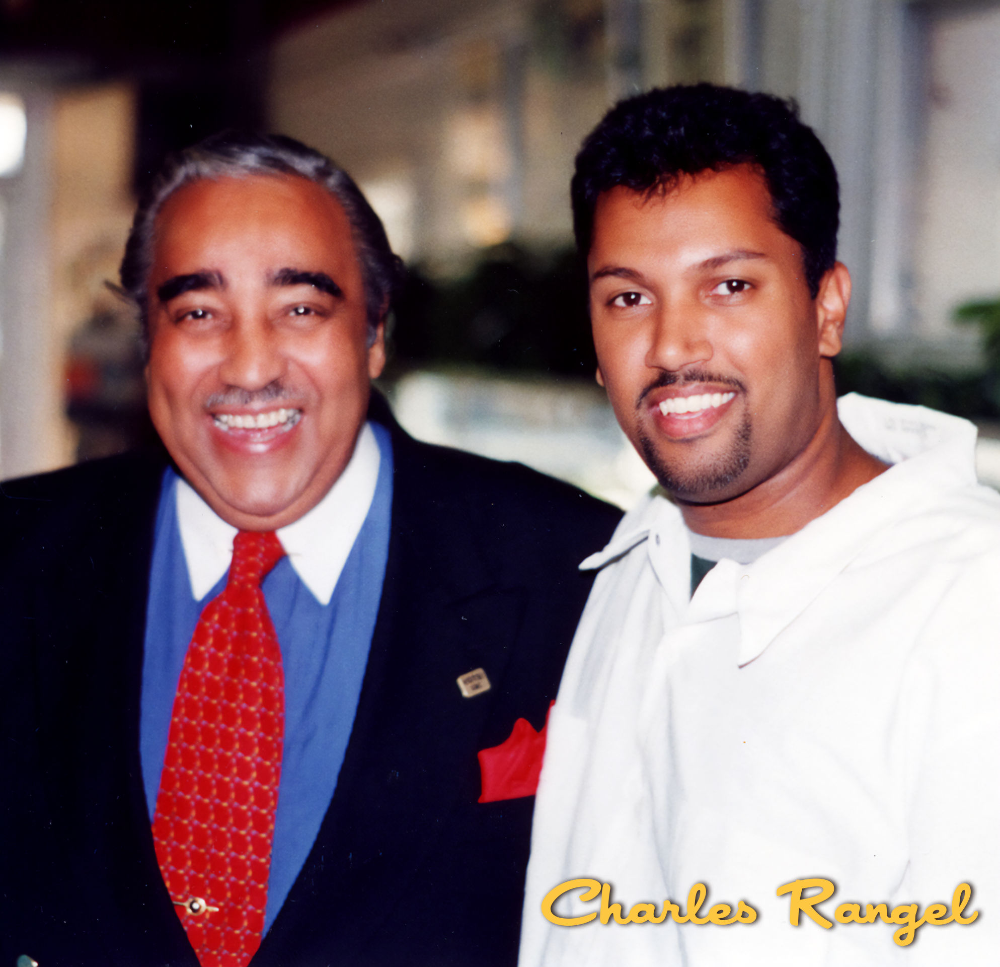 A smiling older man in a black suit, white shirt, red patterned tie, and red pocket square standing next to a smiling younger man in a white hoodie. The background appears to be an indoor public space. The text 'Charles Rangel' is written in yellow c