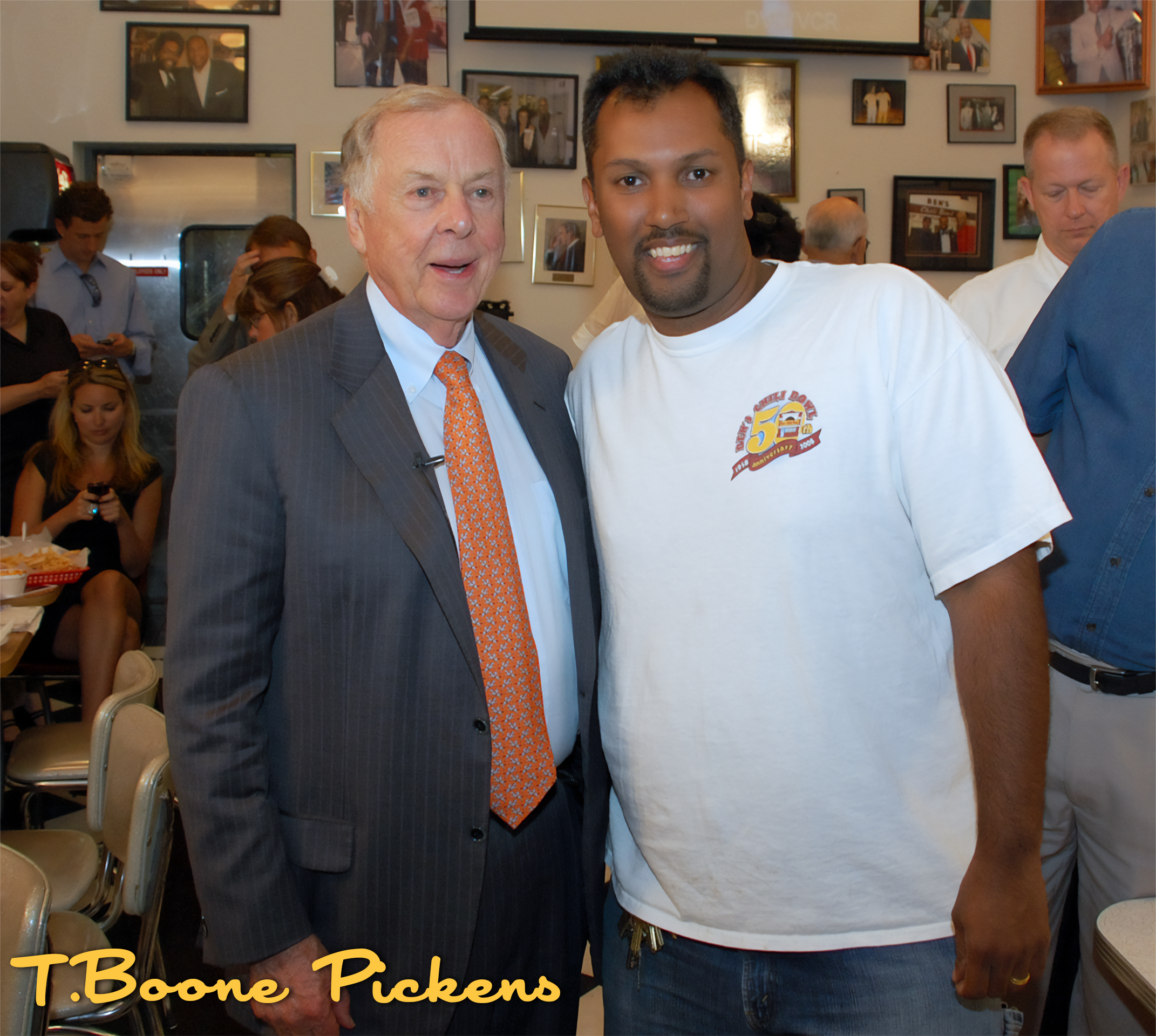 Two men standing and smiling at an event, with a crowded background of people and photos on the wall.