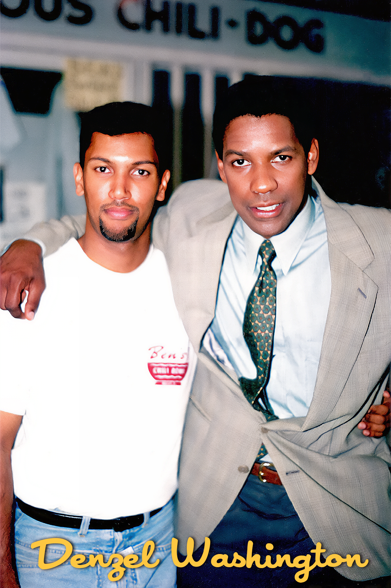 Two men stand side by side with their arms around each other, smiling at the camera. One man is wearing a white t-shirt and the other man is dressed in a suit with a patterned tie. The background is blurred, and the text 'Denzel Washington' is writte