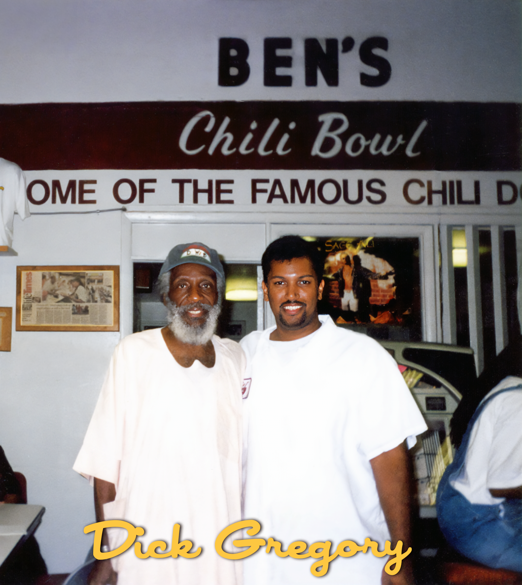 Two men standing inside Ben's Chili Bowl restaurant, smiling at the camera. Behind them, a sign reads "Ben's Chili Bowl," and there is a television and some framed items on the wall. The man on the left has a gray beard and is wearing a t-shirt and c