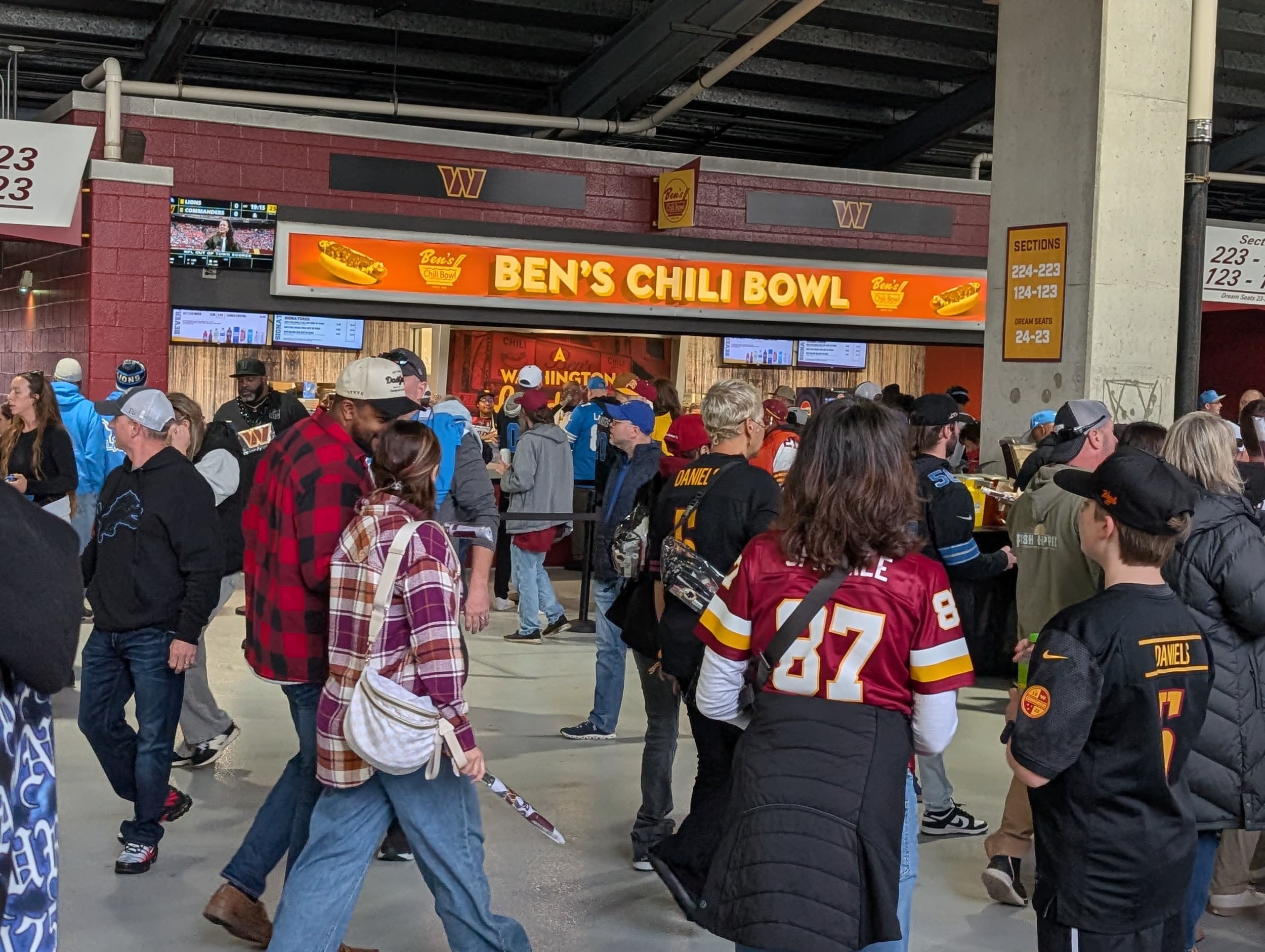 Fans entering Ben's Chili Bowl at a sports stadium, with some wearing football jerseys and others in casual clothing.