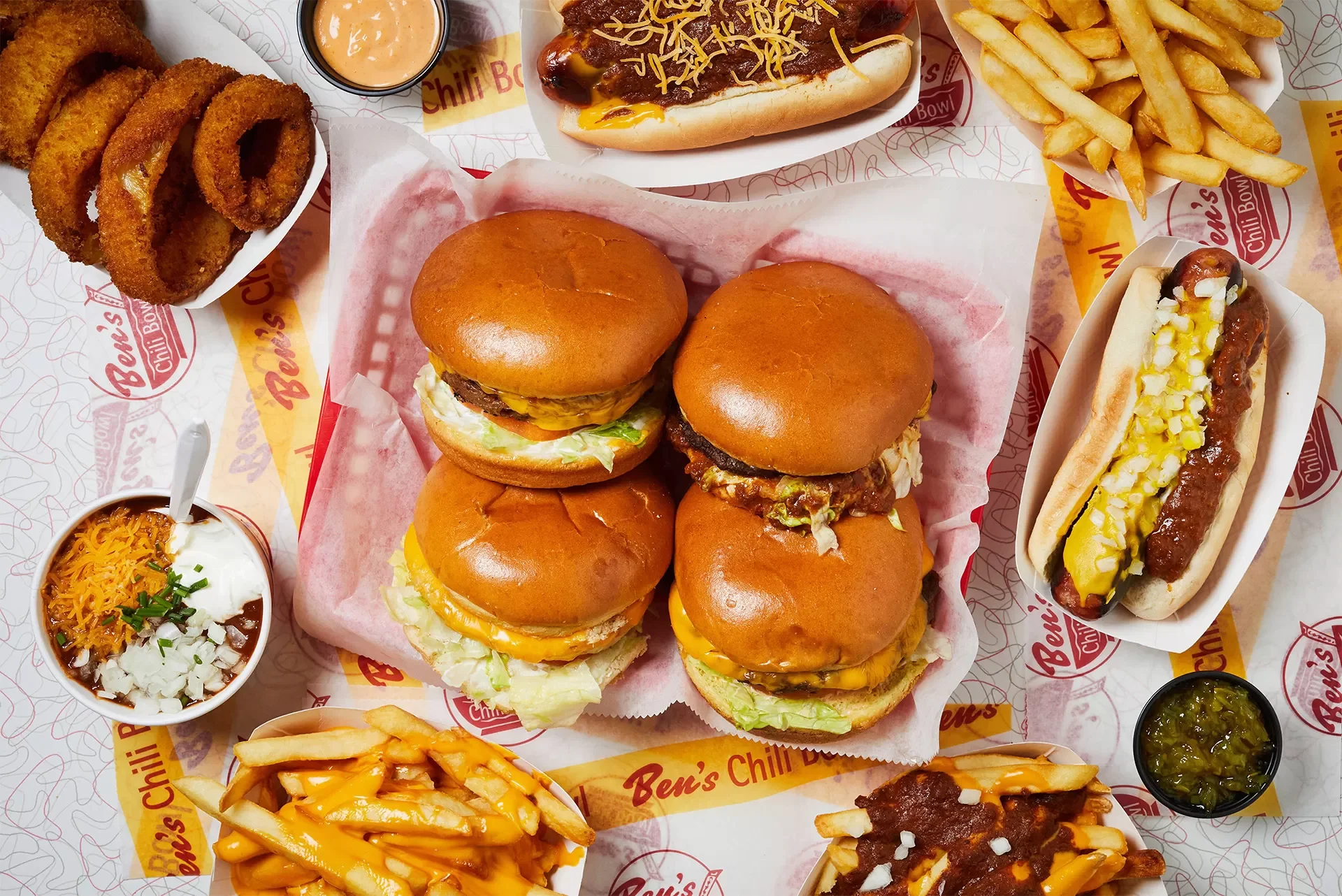 Assorted fast food including four cheeseburgers, a hot dog with mustard and relish, French fries, onion rings, chili cheese fries, a small bowl of chili, and cups of dipping sauces on a patterned paper with Ben's Chili Bowl branding.
