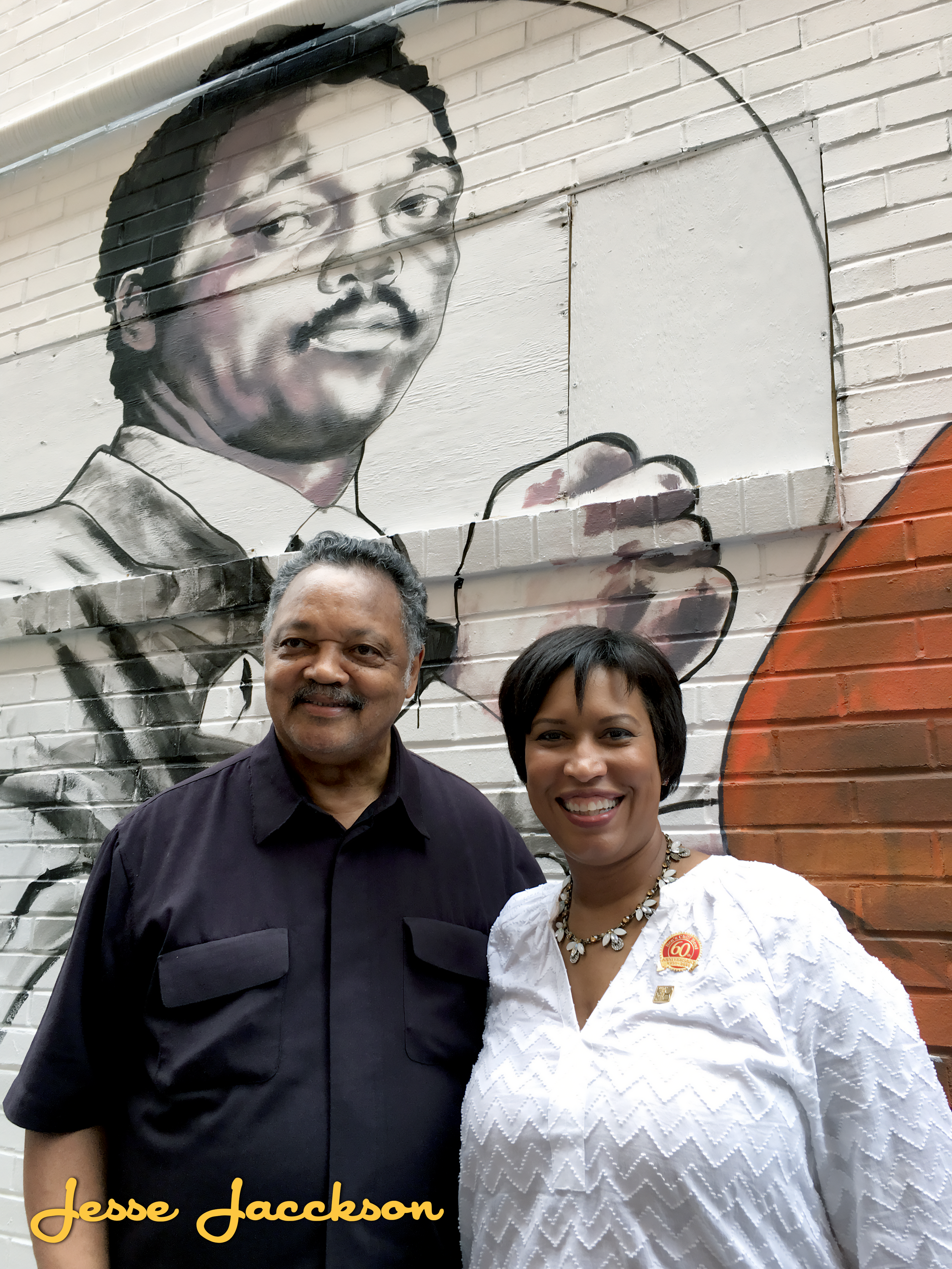 Two people, a man and a woman, standing in front of a mural of a man holding a microphone. The mural is black and white, and the man in the photo is smiling at the camera. The woman is wearing a white blouse and a necklace, and the man is wearing a b