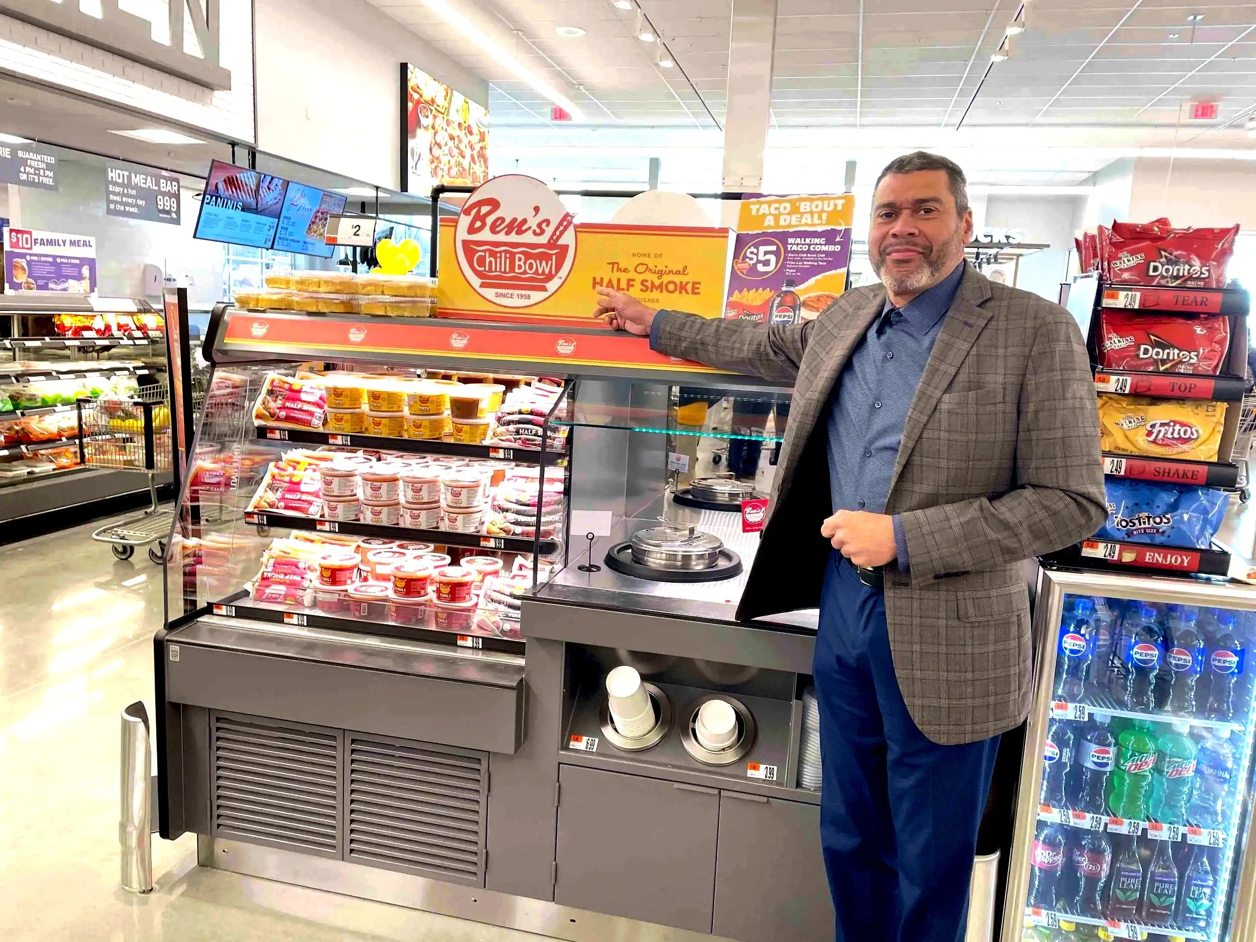 A man in a plaid jacket points to a Ben's Chili Bowl sign at a grocery store. He is standing next to a refrigerated display of yogurt and a hot food counter surrounded by snack shelves and soda cooler.