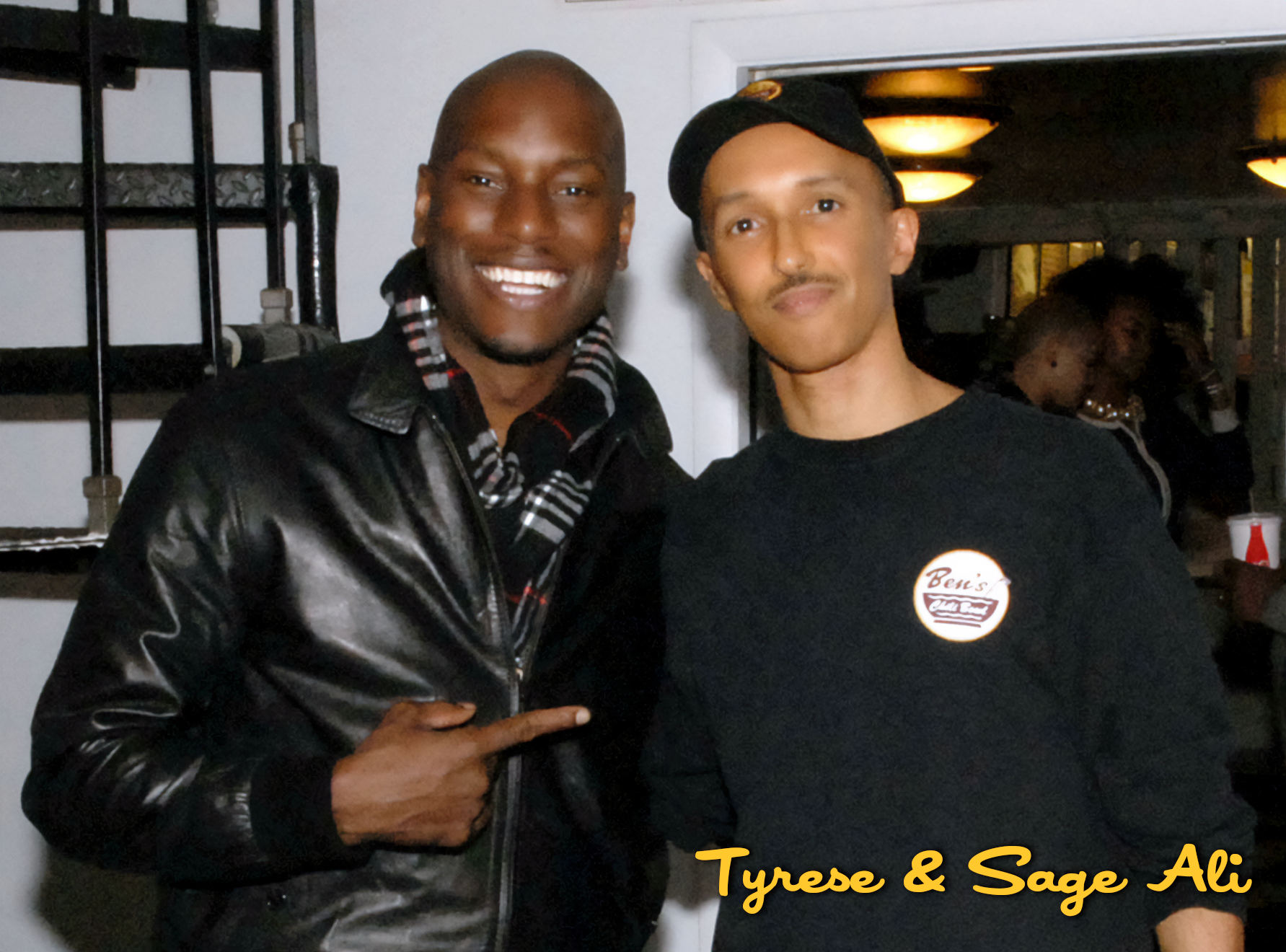 Two men smiling and posing for a photo indoors. The man on the left is wearing a black leather jacket and a striped scarf, while the man on the right is wearing a black hoodie with a logo and a cap. The image has the caption "Tyrese & Sage Ali."