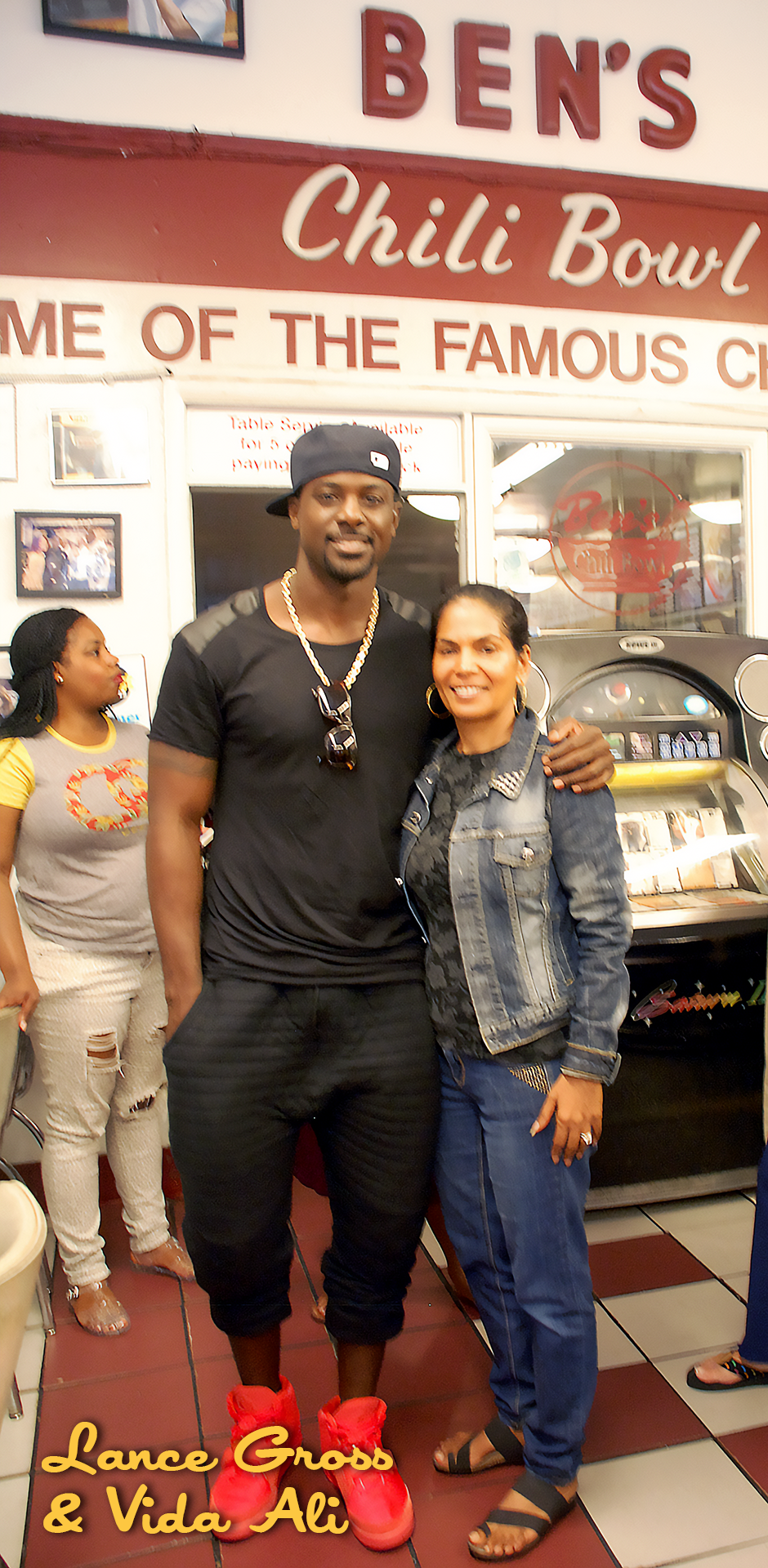 A man and a woman standing together inside a restaurant named Ben's Chili Bowl, with the man wearing a black cap, black T-shirt, and red shoes, and the woman wearing a denim jacket and blue jeans. There is a young girl in the background wearing a gra
