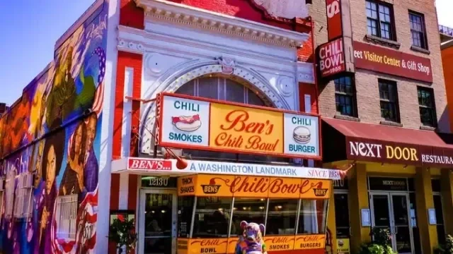 A street scene showing the facade of Ben's Chili Bowl, a historic restaurant with colorful signage advertising chili, smoked, burgers, and the restaurant's name. There is a small food cart in front with similar signage. Buildings and signs for other businesses are visible in the background.
