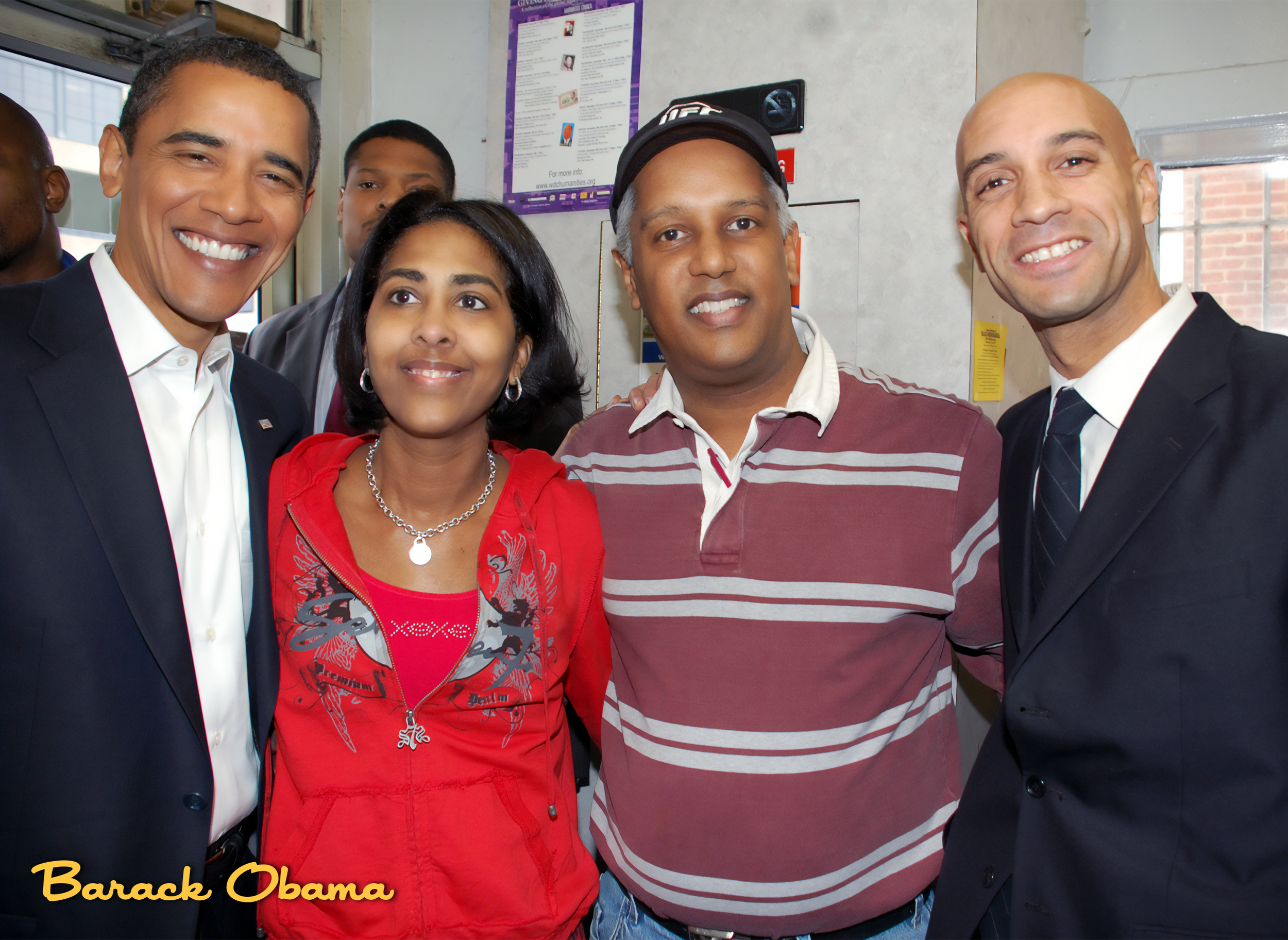 Group of five diverse people smiling for a photo indoors, with two men in suits, a woman in a red jacket, and two other men. The bottom left corner has the name "Barack Obama" written in yellow text.