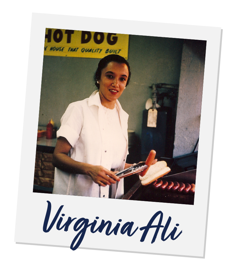 A woman in a white shirt cooking hot dogs on a grill with a bun nearby, standing in front of a yellow sign that says 'HOT DOG' in the background.