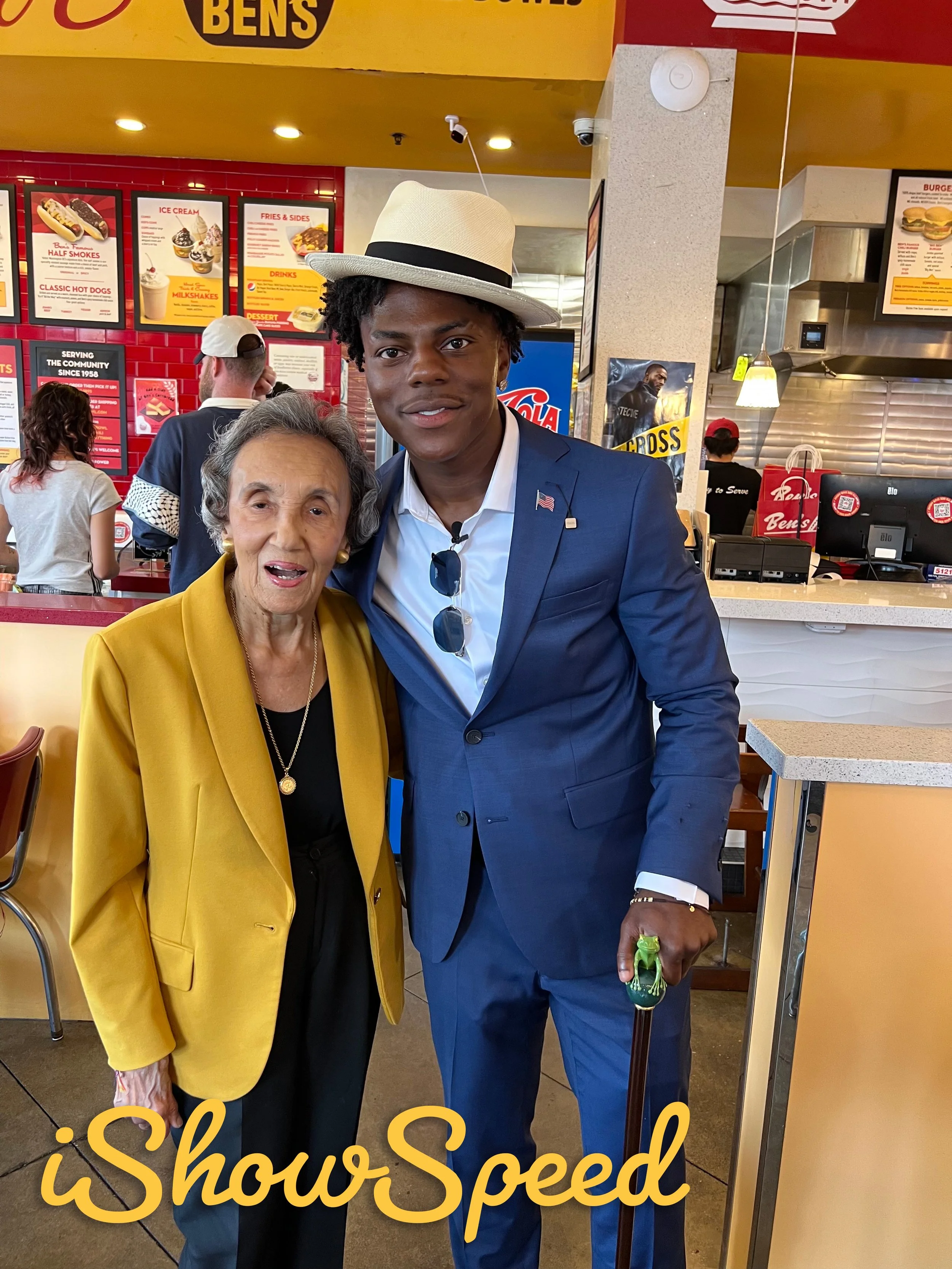 A young man in a blue suit and hat standing inside a fast food restaurant, posing with an elderly woman in a yellow blazer. The background features a menu board, counter area, and other customers.