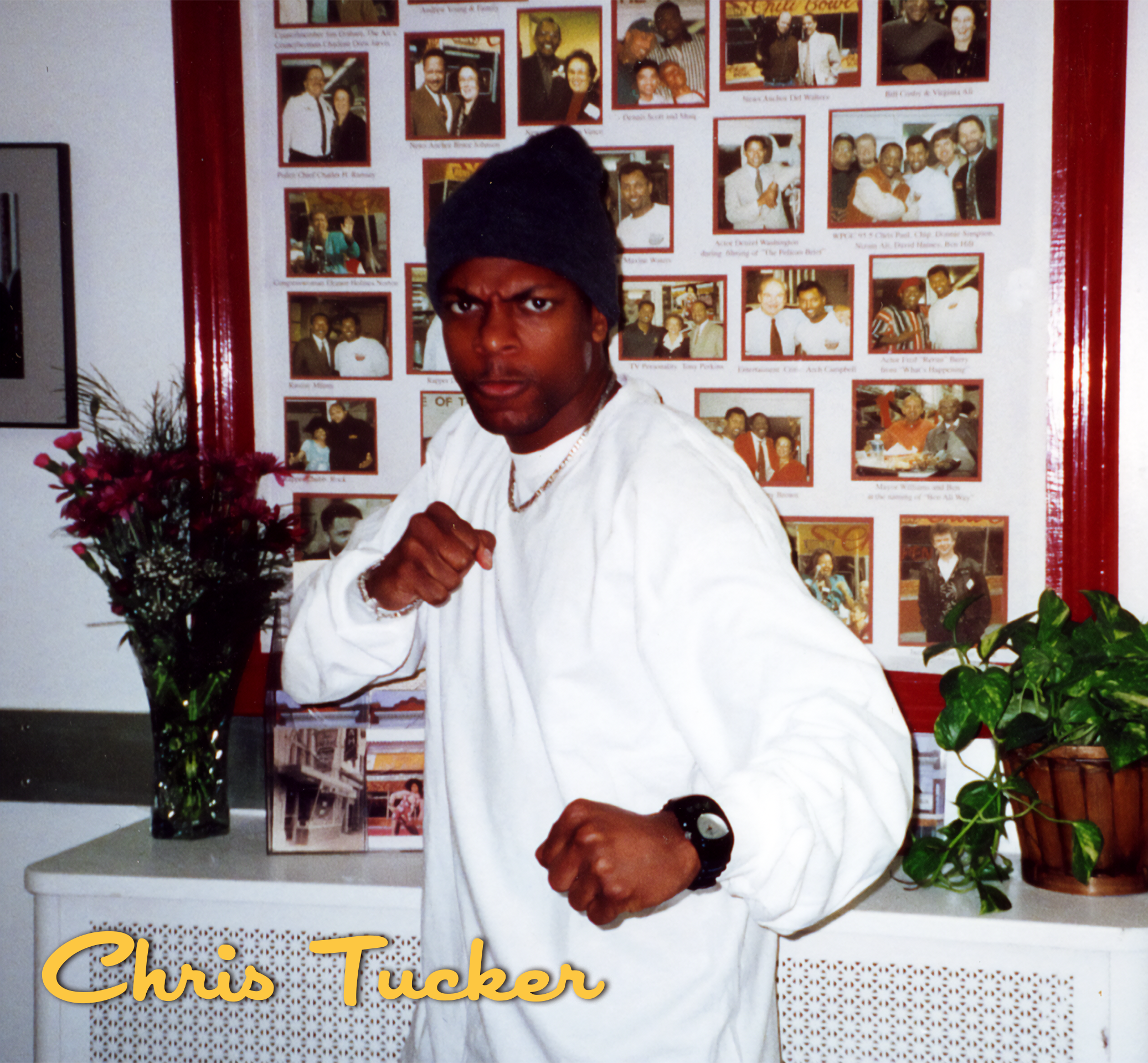 A young man with a serious expression wearing a black hat and white sweatshirt, holding a fist up in a boxing stance, with a collage of photographs on a board behind him, and a name label reading "Chris Tucker" in yellow text at the bottom left corne